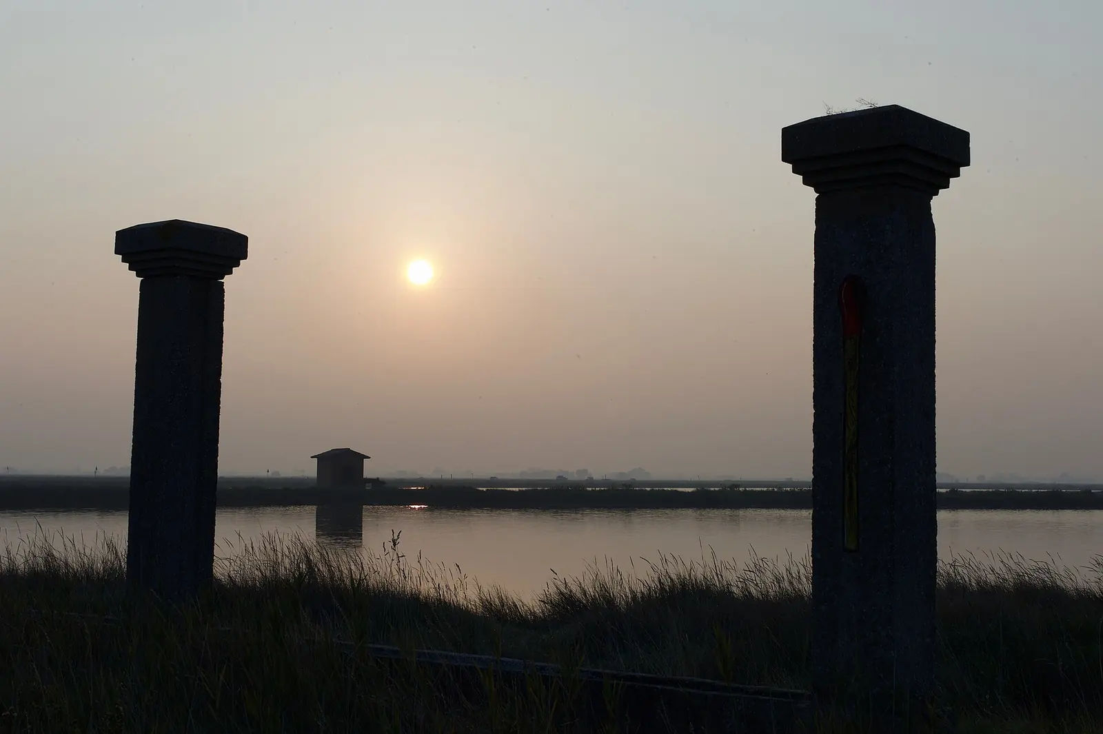 Zwei Pfeiler als Silhouetten vor einer Wasserfläche der Salina Camillone, mit von Dunst verschleierter Sonne