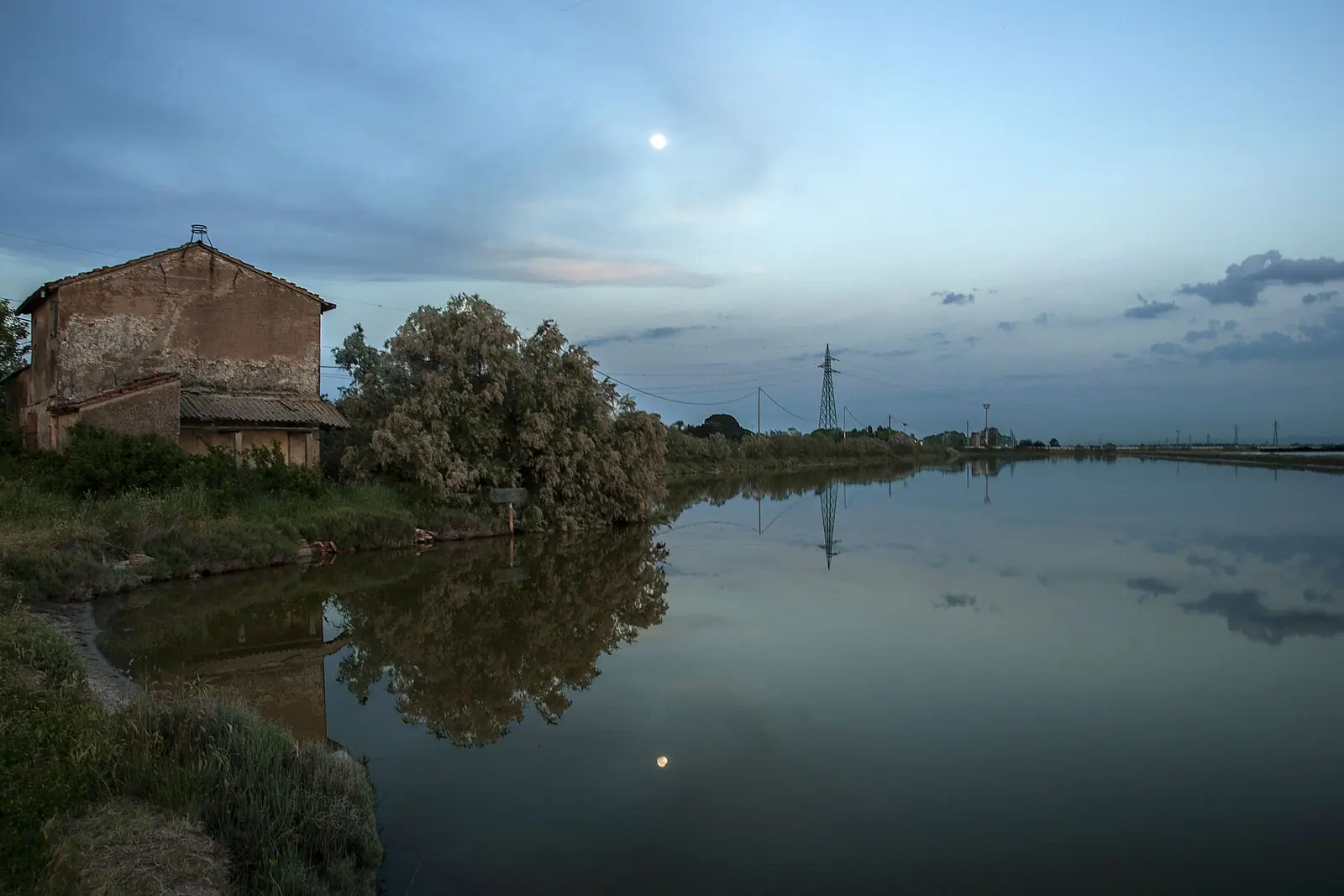 Ein Kanal der Saline von Cervia in der Dämmerung mit einem alten Bauernhaus, einem blühenden Strauch und dem Mond