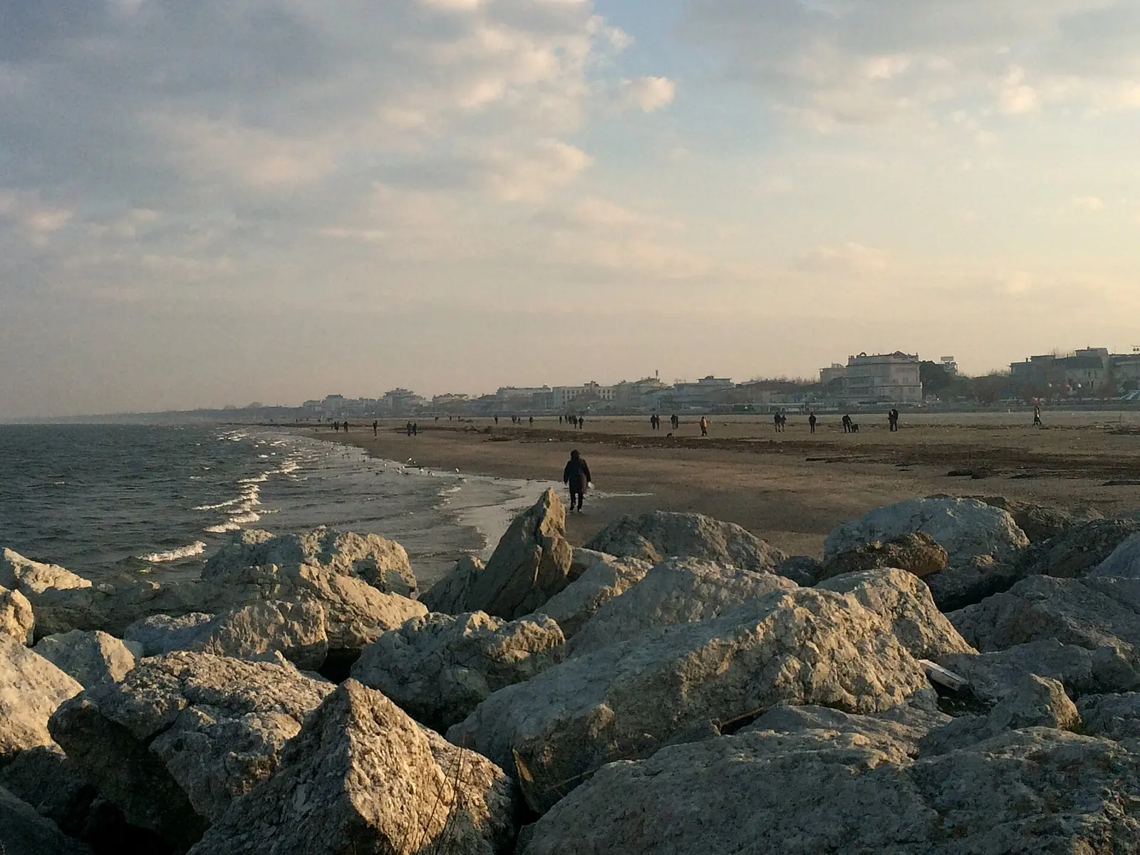 La spiaggia di Cervia in inverno vista dagli scogli, con alcune persone a passeggio lungo la riva e i palazzi lontani