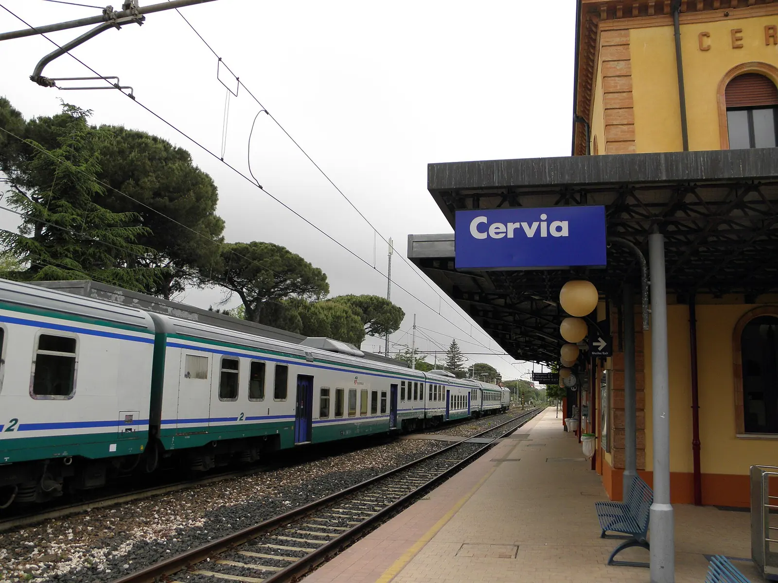 A train at a platform at Cervia-Milano Marittima station, with the canopy and pines in the background