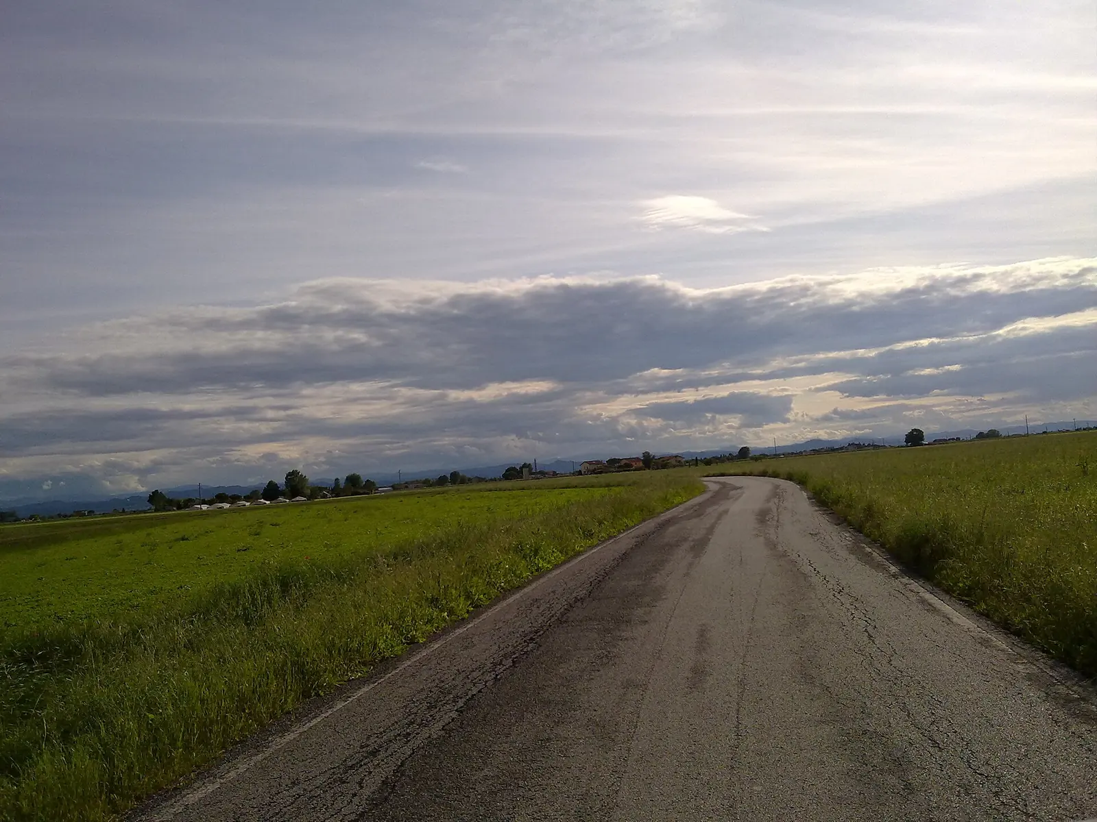 A paved road through green fields near Tagliata, beneath a cloudy sky
