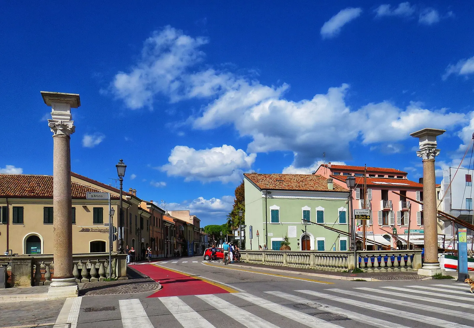 Die beiden Steinsäulen an der Einfahrt des Kanalhafens von Cesenatico mit Straße und bunten Häusern hinter der Balustrade