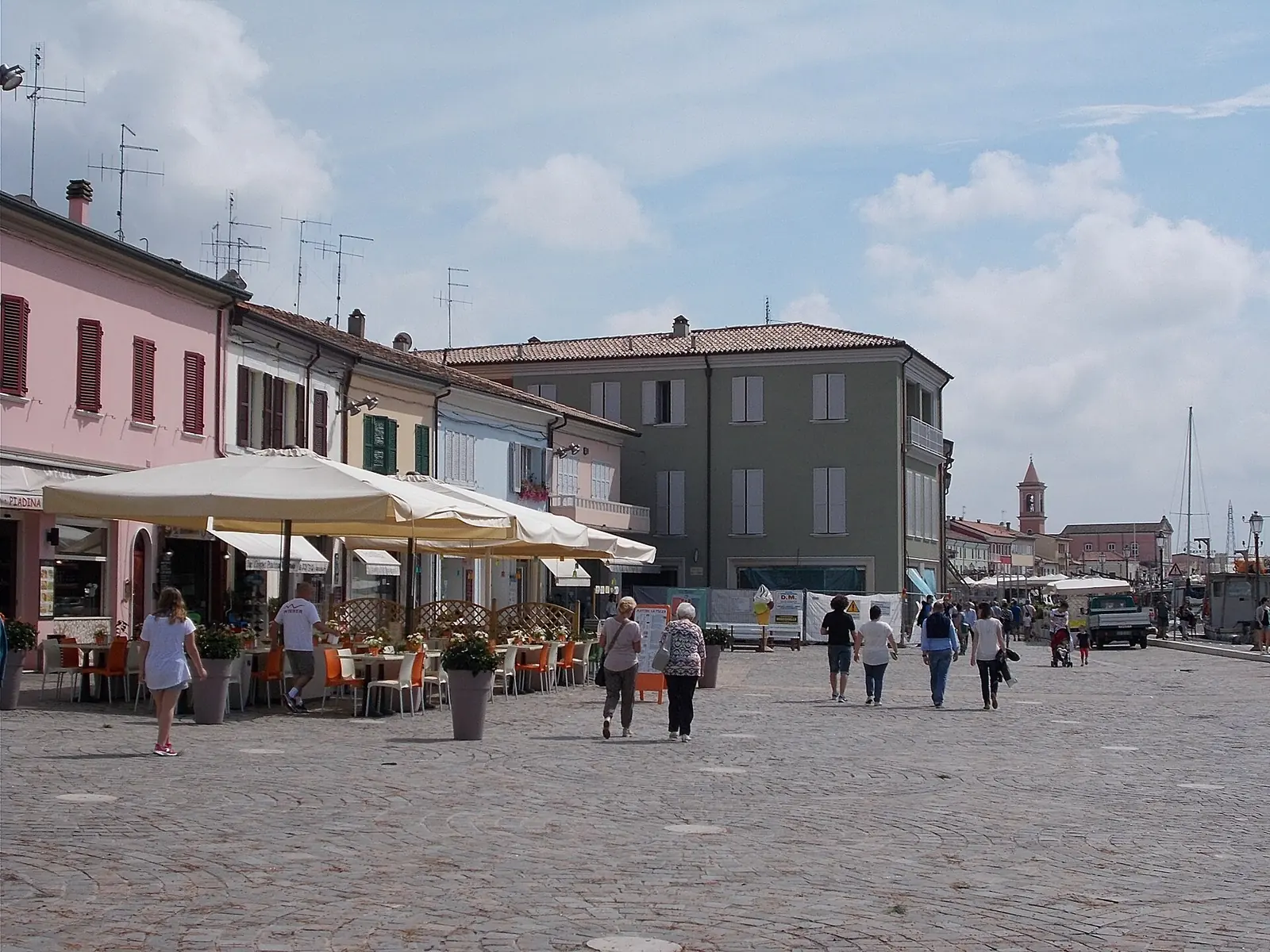 Ein gepflasterter Platz im Zentrum von Cesenatico mit Tischen im Freien unter großen Sonnenschirmen und einigen Spaziergängern