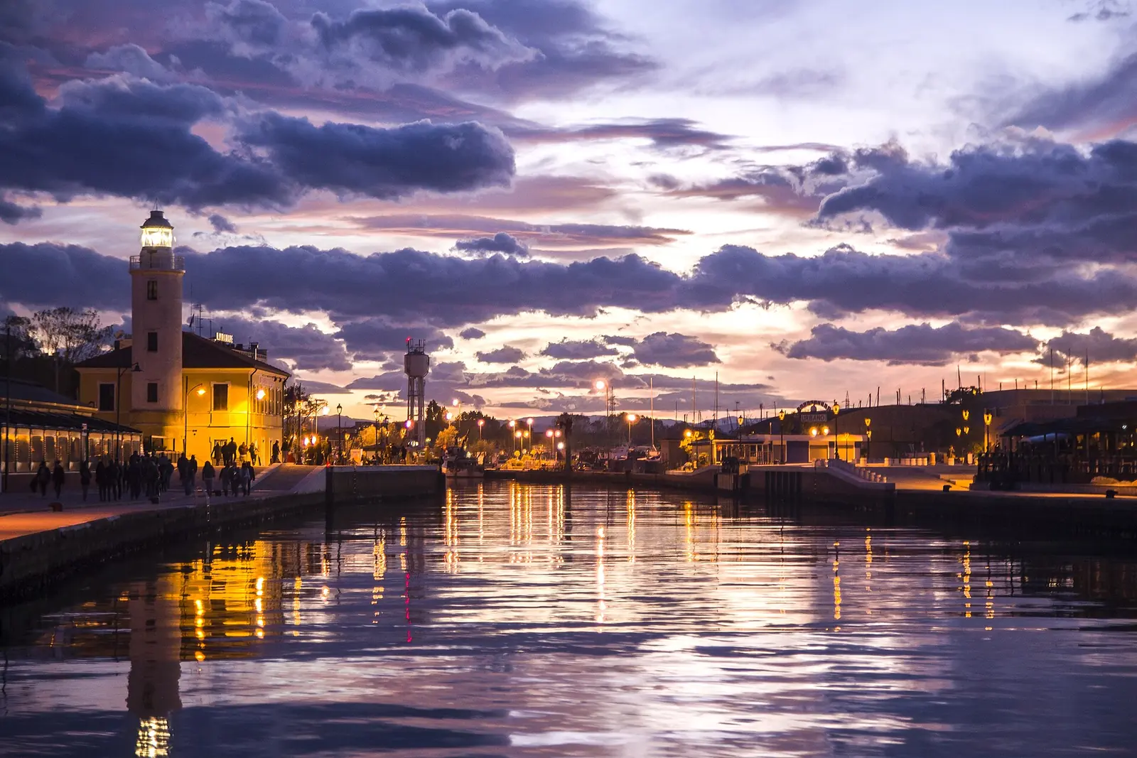 Der Leuchtturm und der Kai des Kanalhafens von Cesenatico bei Sonnenuntergang mit violetten Wolken und eingeschalteten Laternen