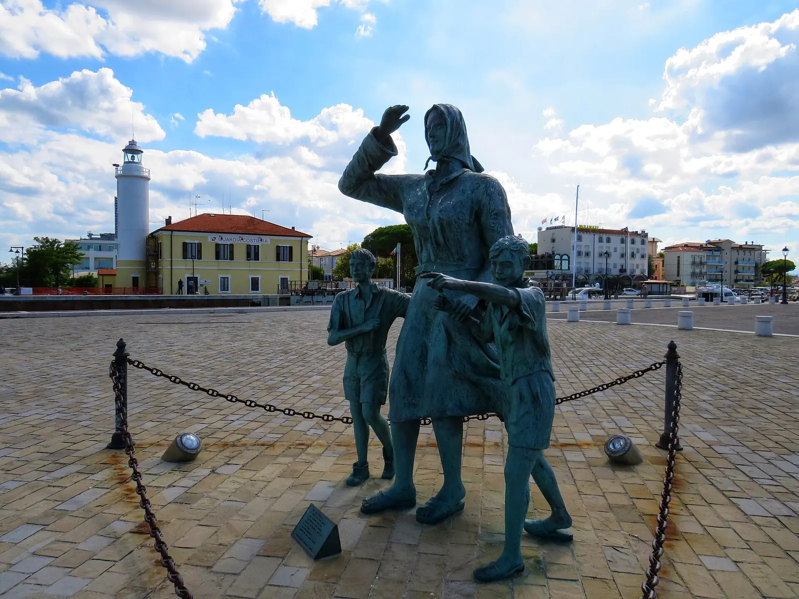 Die Bronzegruppe einer Frau mit zwei Kindern beim Blick auf das Meer an der Piazza Spose dei Marinai mit dem Leuchtturm dahinter