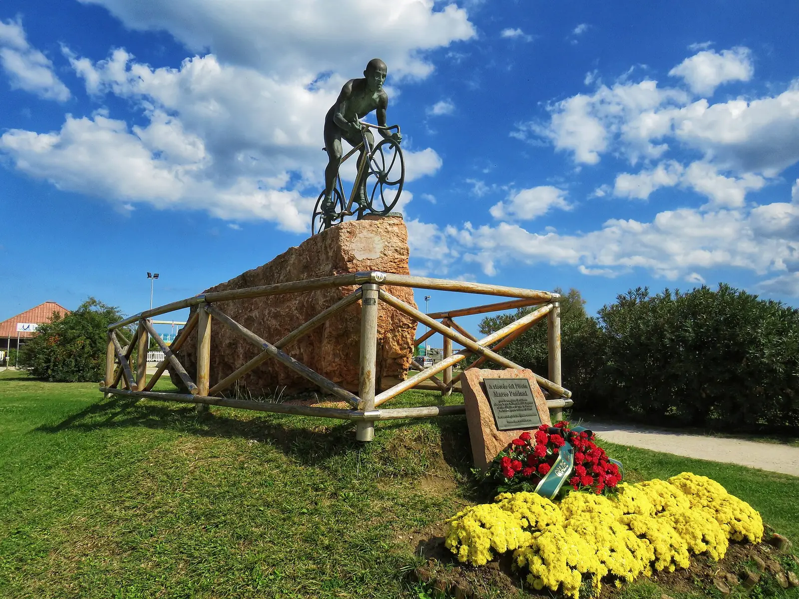 Das Bronzedenkmal eines Radfahrers auf einem Steinblock auf einer Wiese mit Blumen und einem Holzzaun