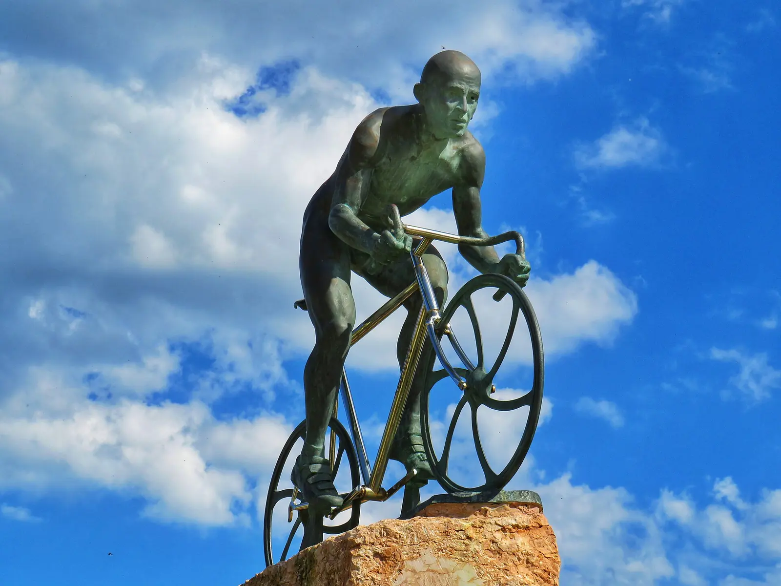 Die Bronzestatue eines Radfahrers beim Anstieg auf einen Felsen, von unten vor blauem Himmel mit Wolken