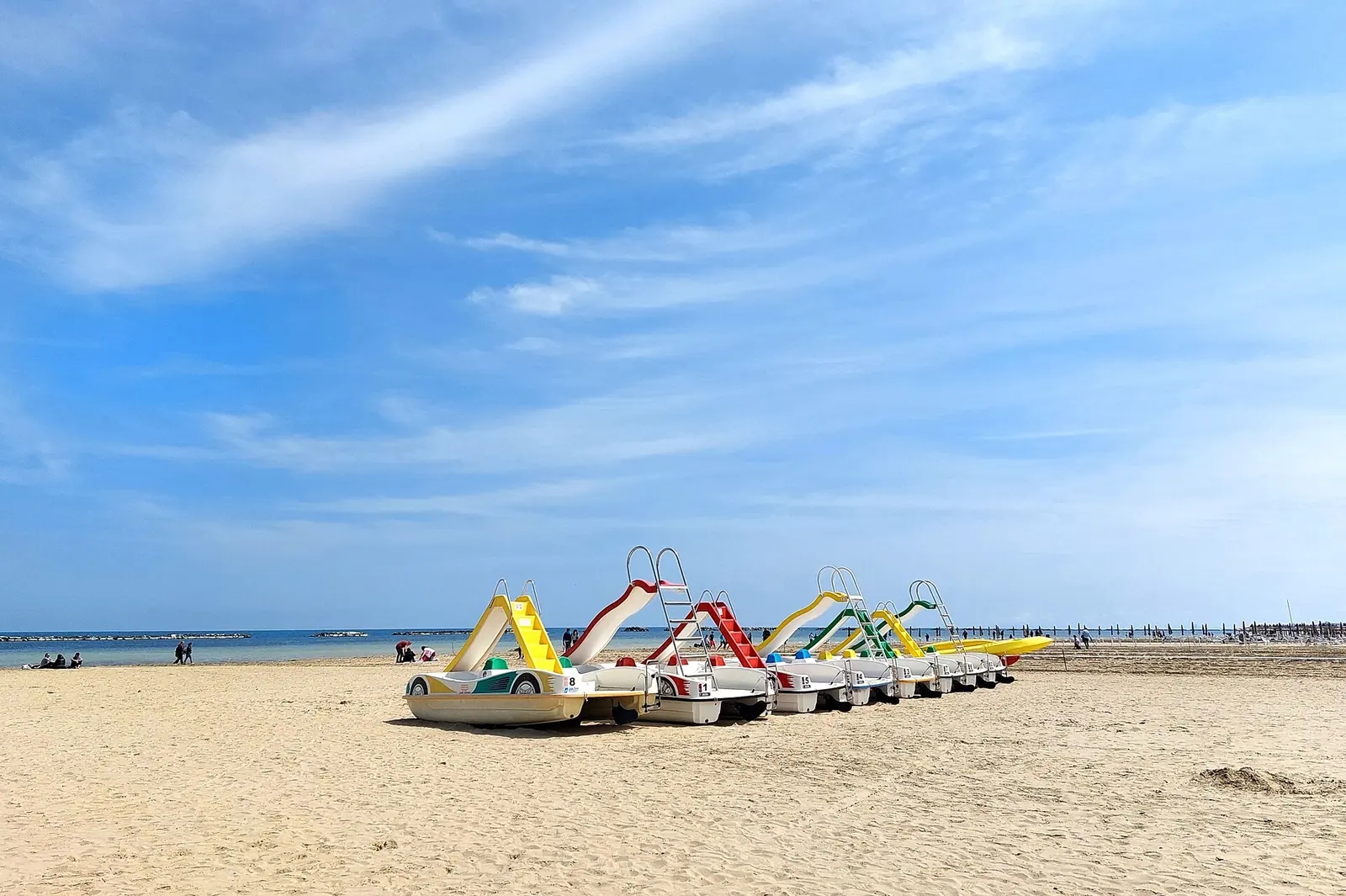 A row of pedal boats with colourful slides lined up on pale sand, with the sea and blue sky behind