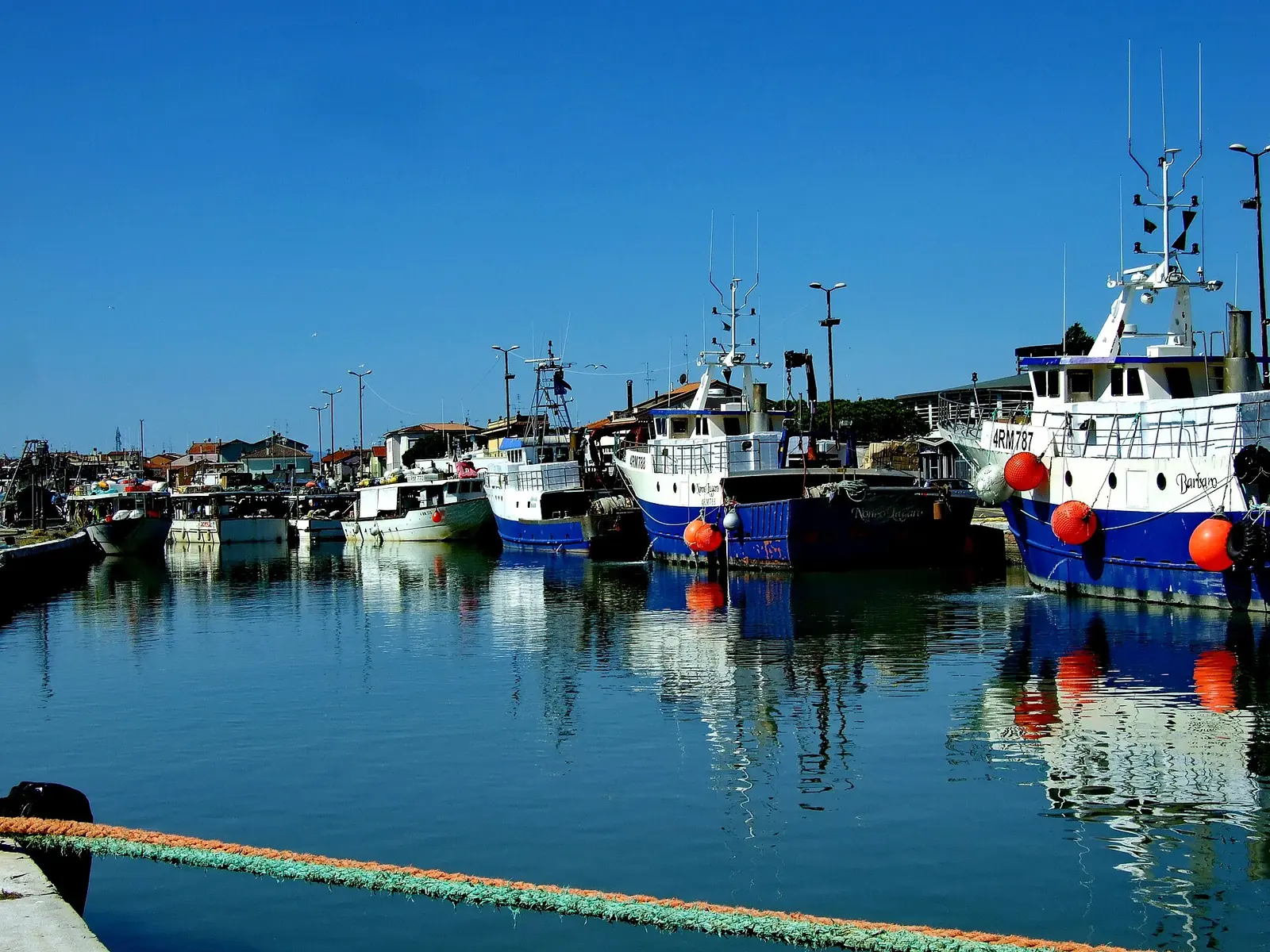 Bateaux de pêche aux bouées orange amarrés dans le port-canal de Cesenatico, avec le ciel bleu et les lampadaires du quai