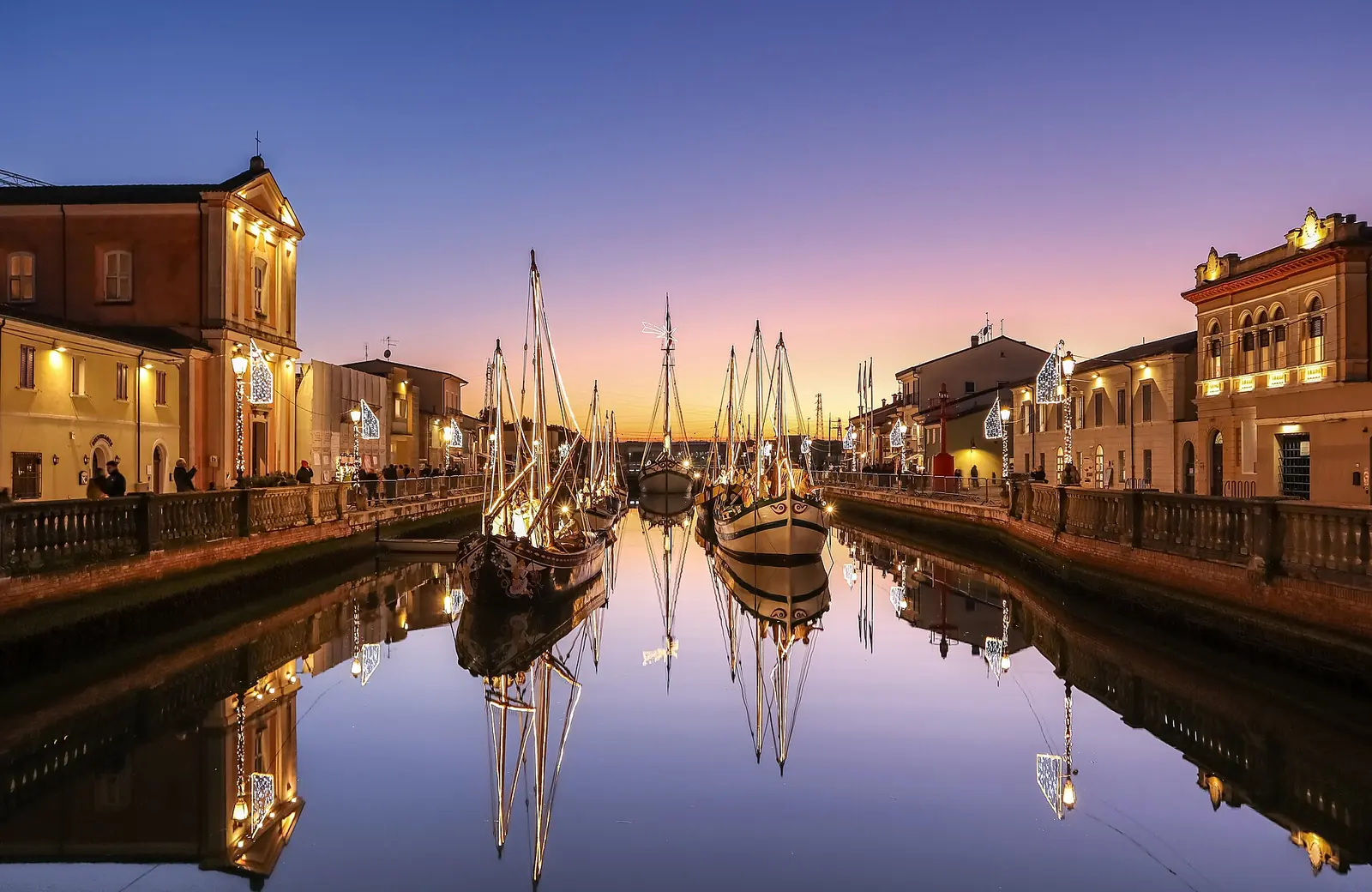 Historic boats in Cesenatico's canal harbour at dusk, with lights on along the quays and a pinkish sky