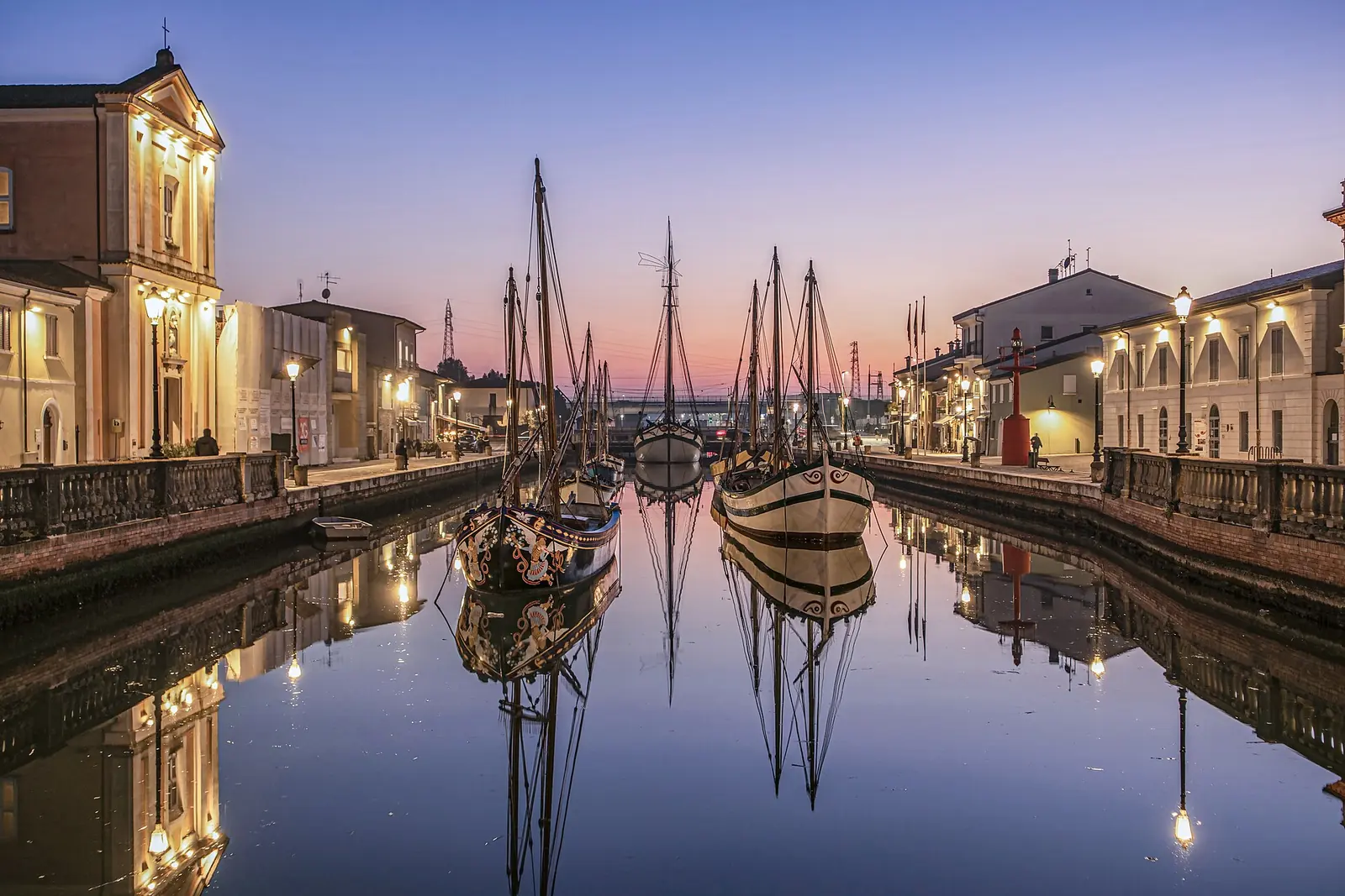 Cesenatico's canal harbour at dusk, with historic boats reflected in the water and lit streetlamps on the quays