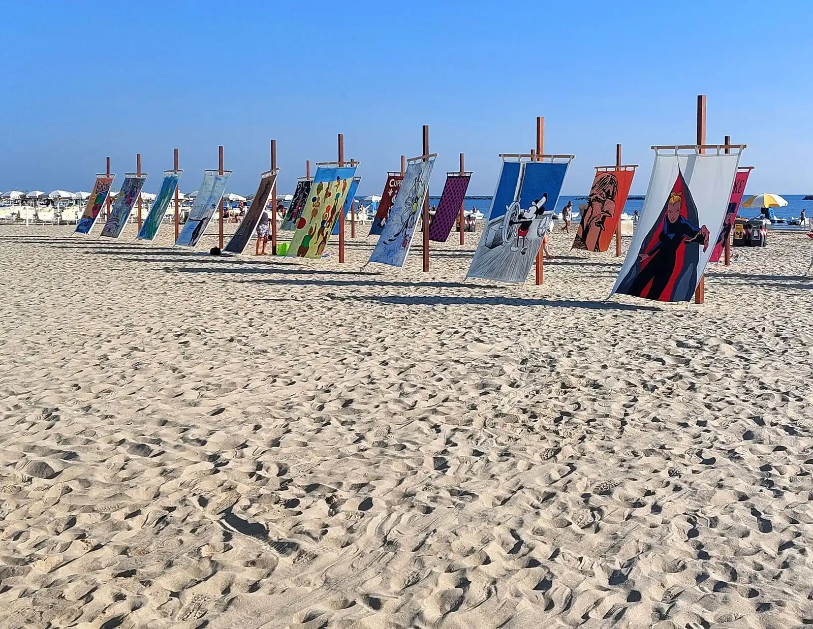 Des toiles peintes suspendues à des poteaux en bois sur une étendue de sable, avec les parasols et la mer en arrière-plan