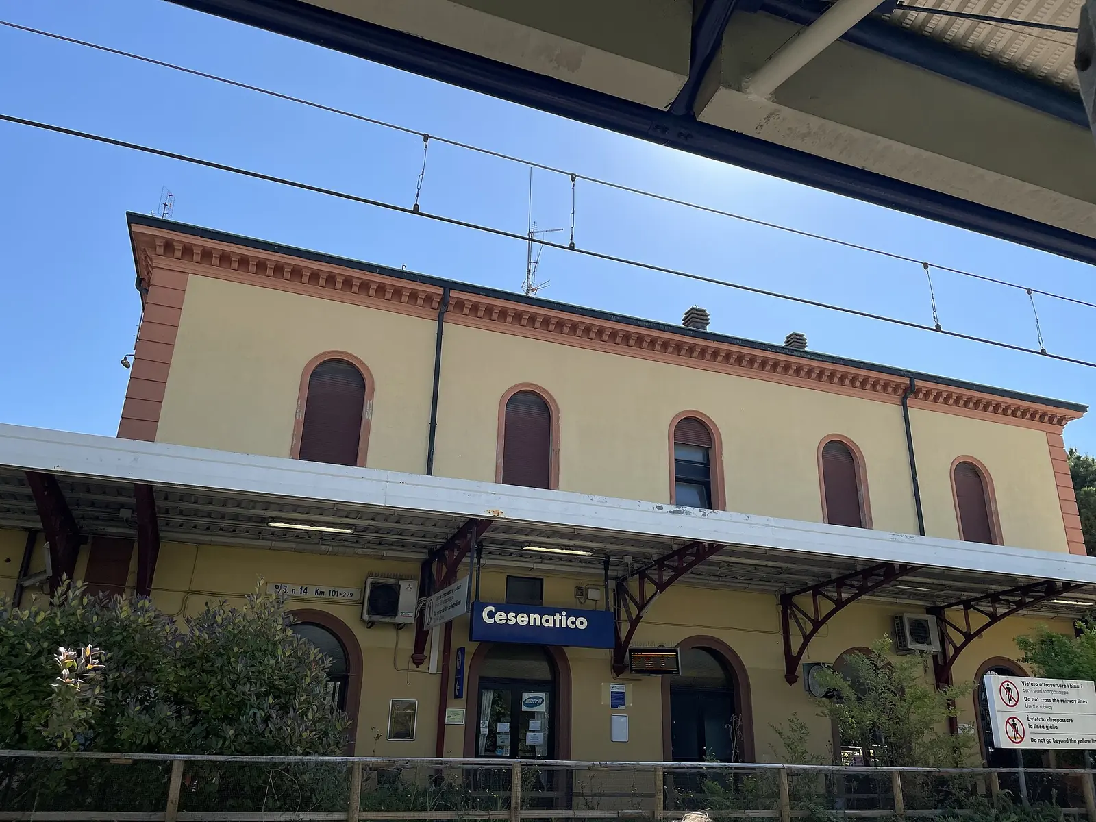 Das gelbe Empfangsgebäude des Bahnhofs Cesenatico vom Gehweg aus mit blauem Schild und Vordach