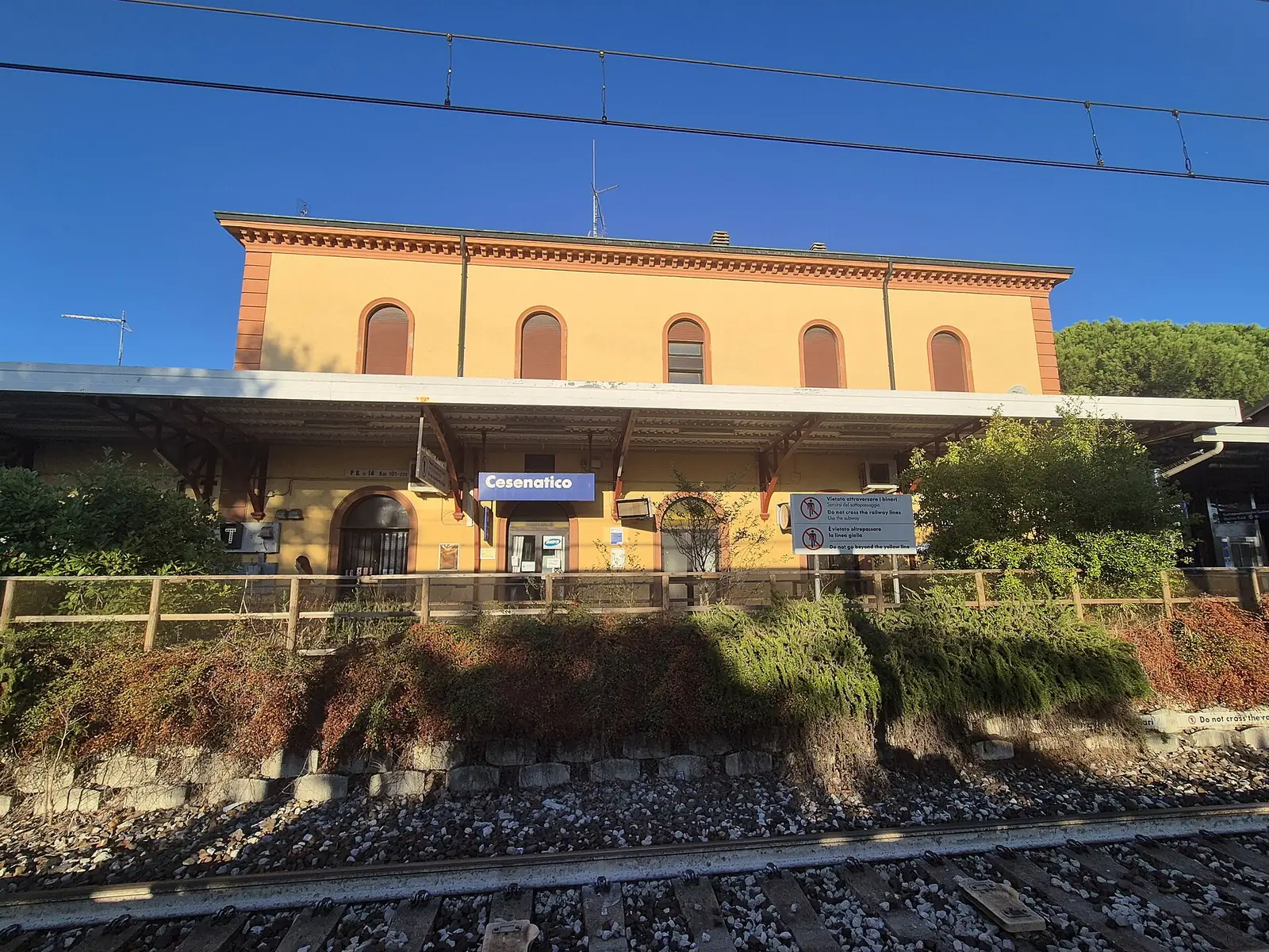 The historic Cesenatico station building seen from the tracks, with overhead wires and the name sign