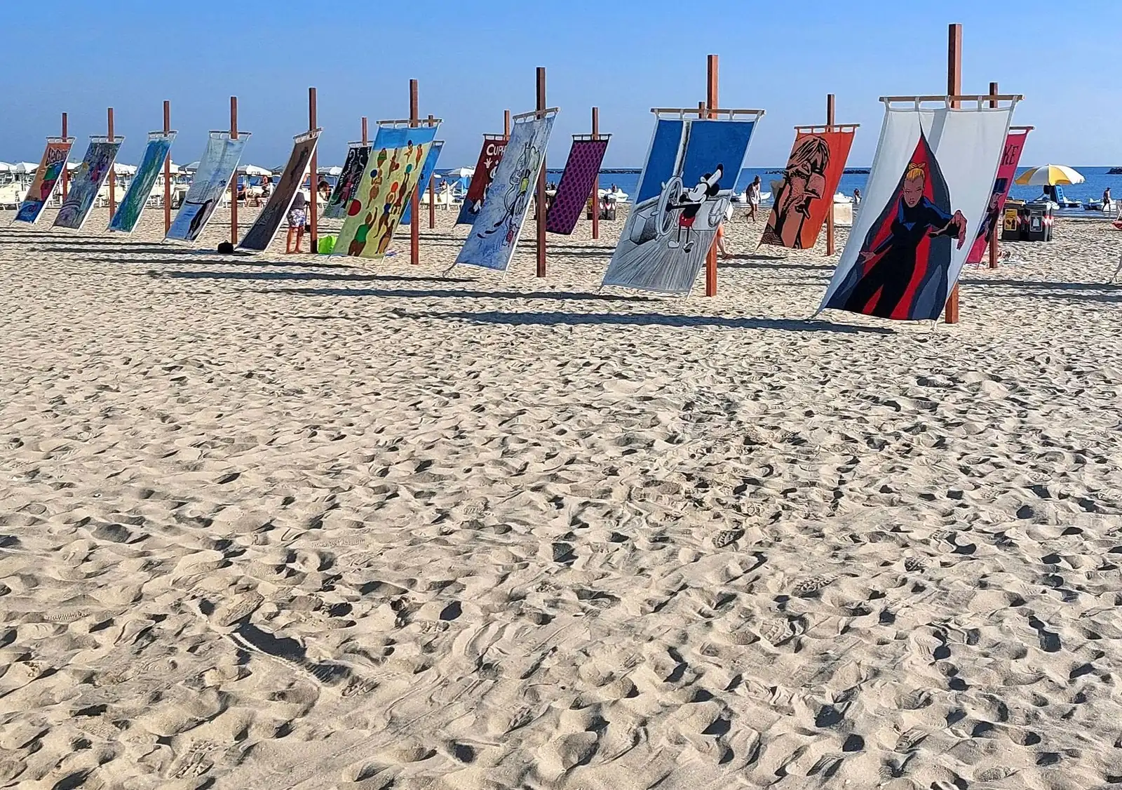 Bemalte Tücher an Holzpfosten im Sand eines freien Strands von Cesenatico, mit dem Meer in der Ferne
