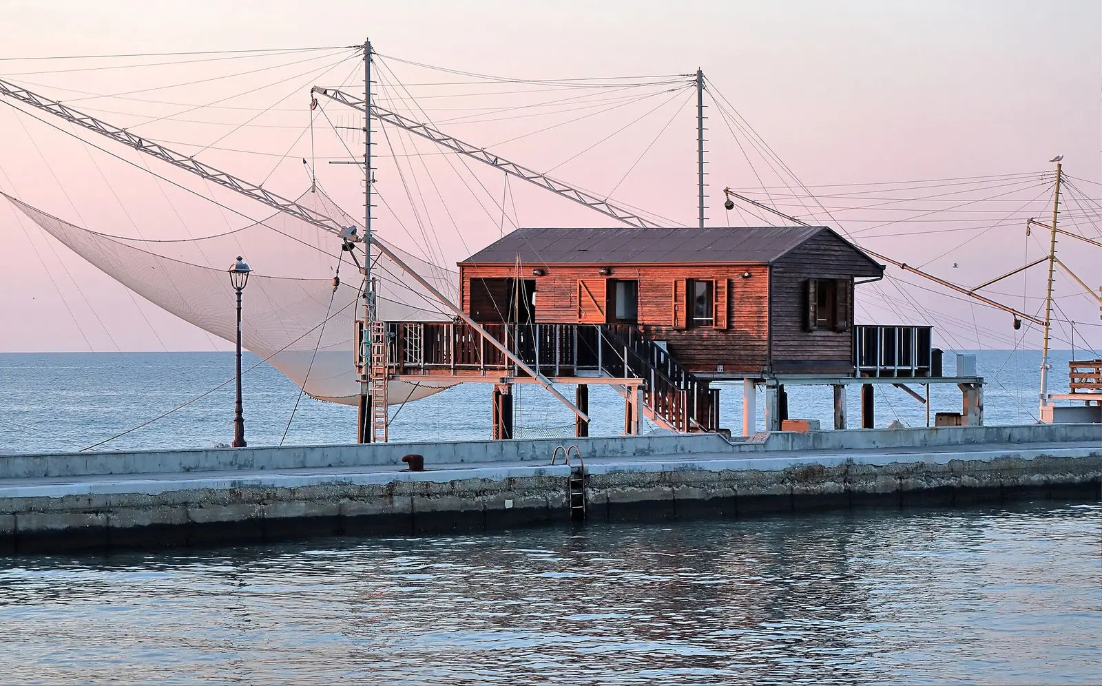 A wooden fishing hut on stilts with its large net raised, on the Cesenatico pier at sunset