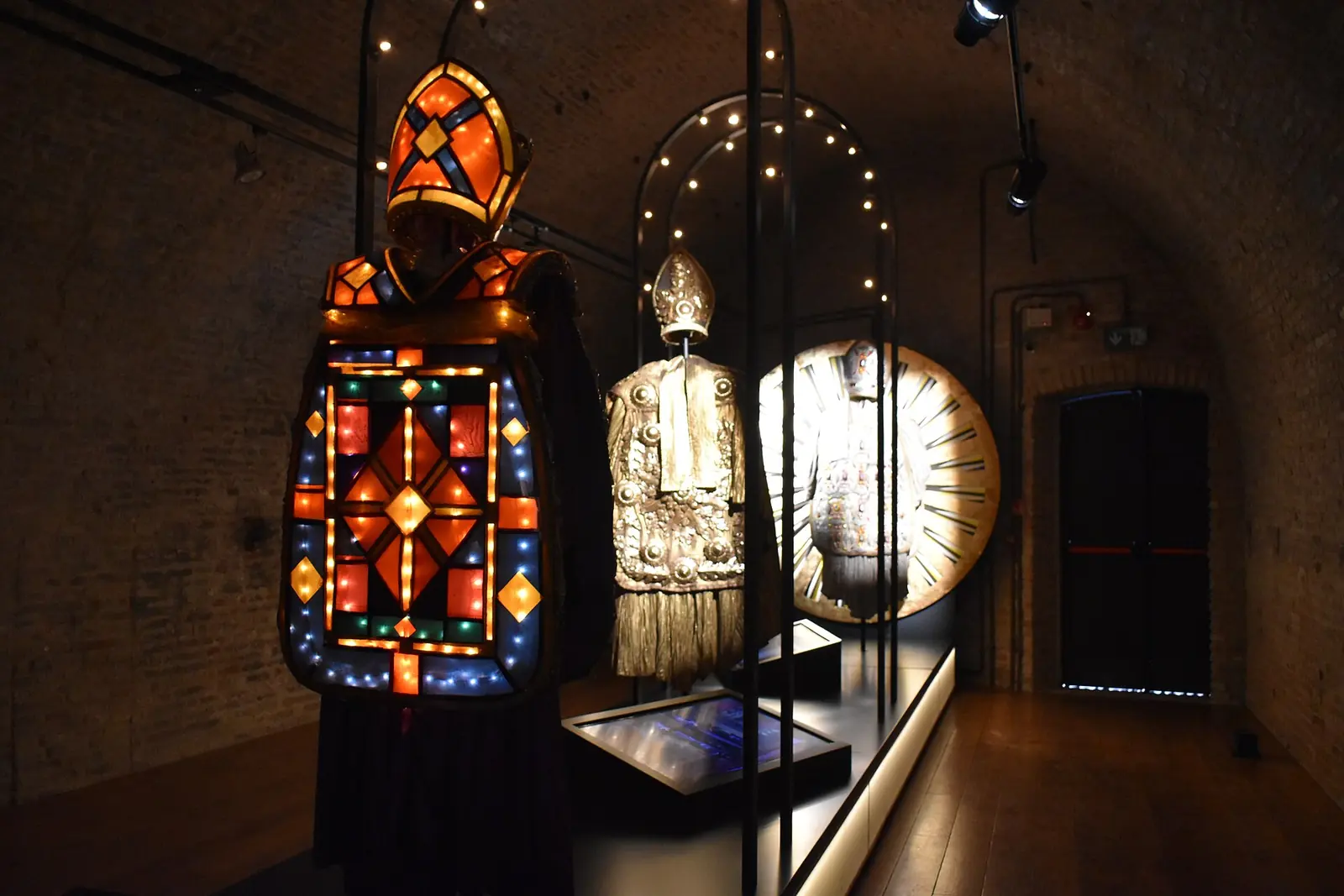Bühnenkostüme mit Mitren und bunten Lichtern in einem Raum mit Backsteingewölbe im Fellini Museum in Rimini