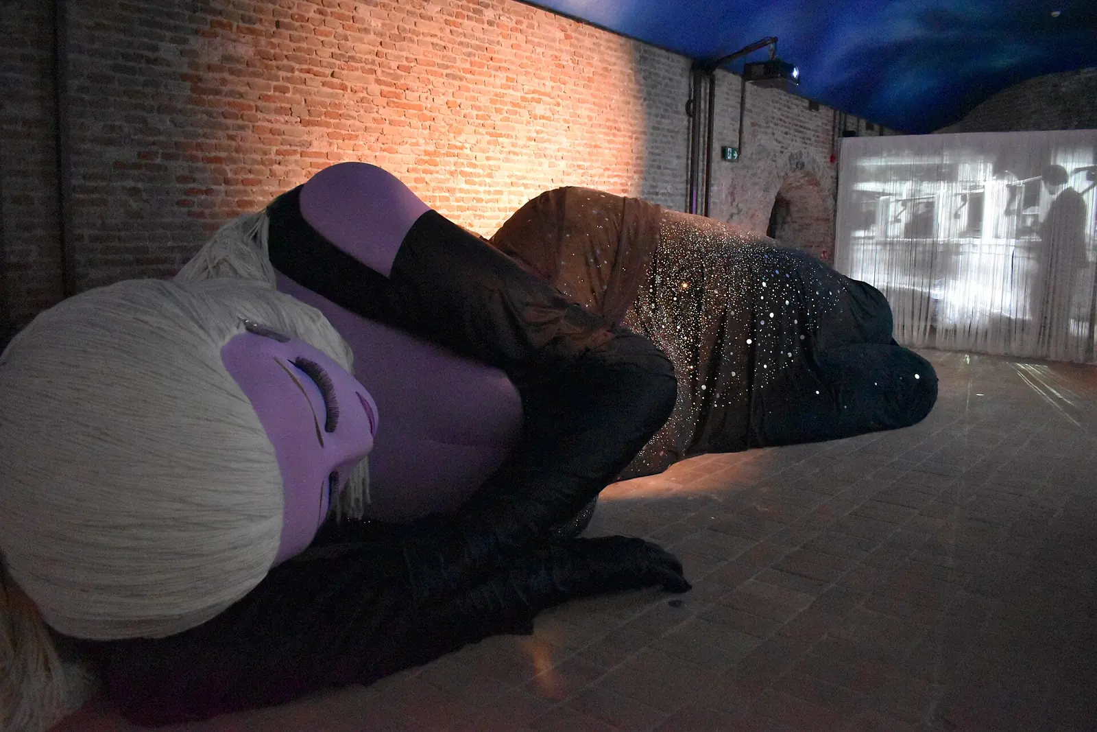 Eine große liegende Figur mit lilafarbenem Gesicht und Wollhaaren in einem Backsteinraum des Fellini Museum in Rimini