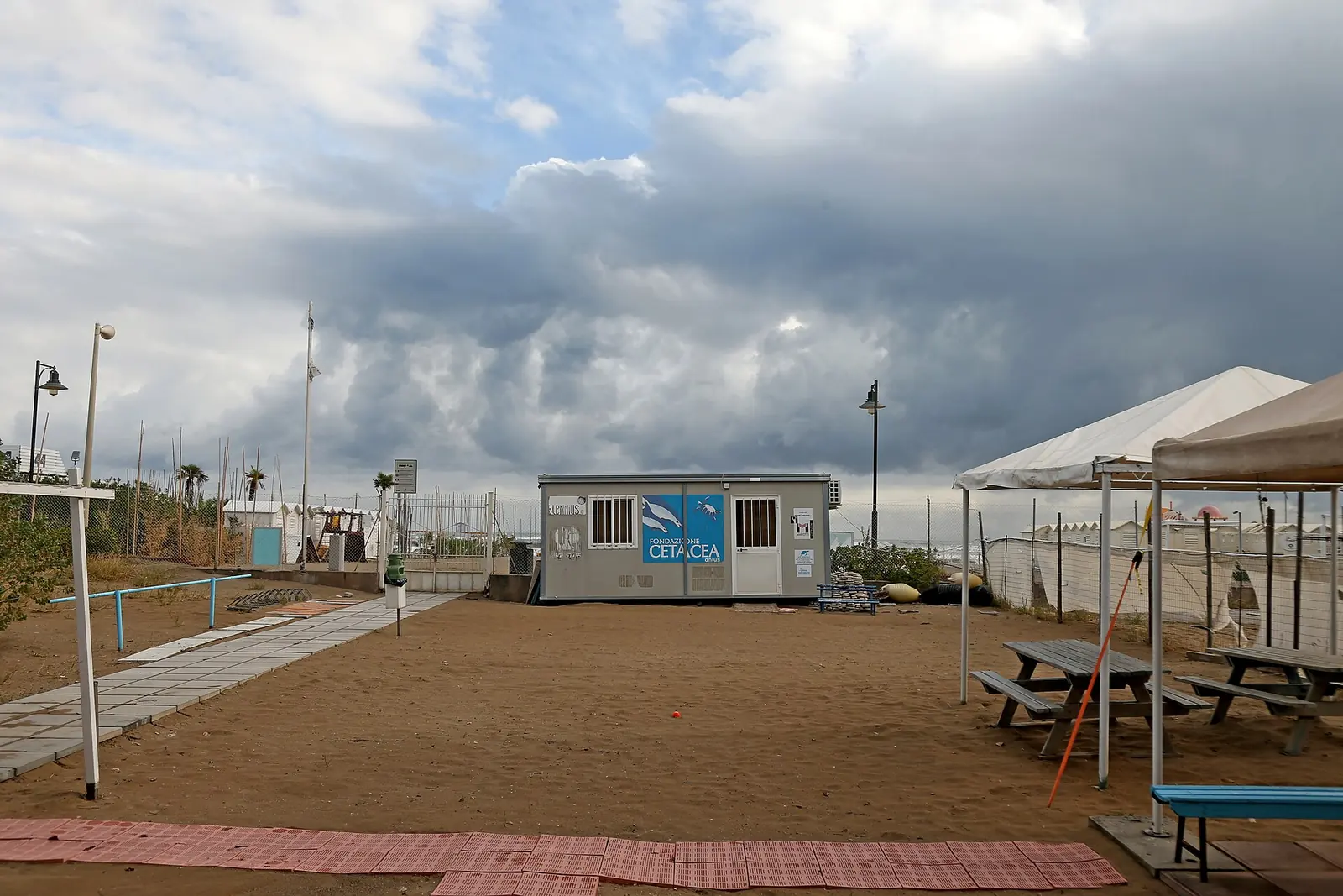 Le bâtiment préfabriqué de la Fondazione Cetacea sur une zone de sable, avec tables de pique-nique et tonnelle sous un ciel chargé de nuages