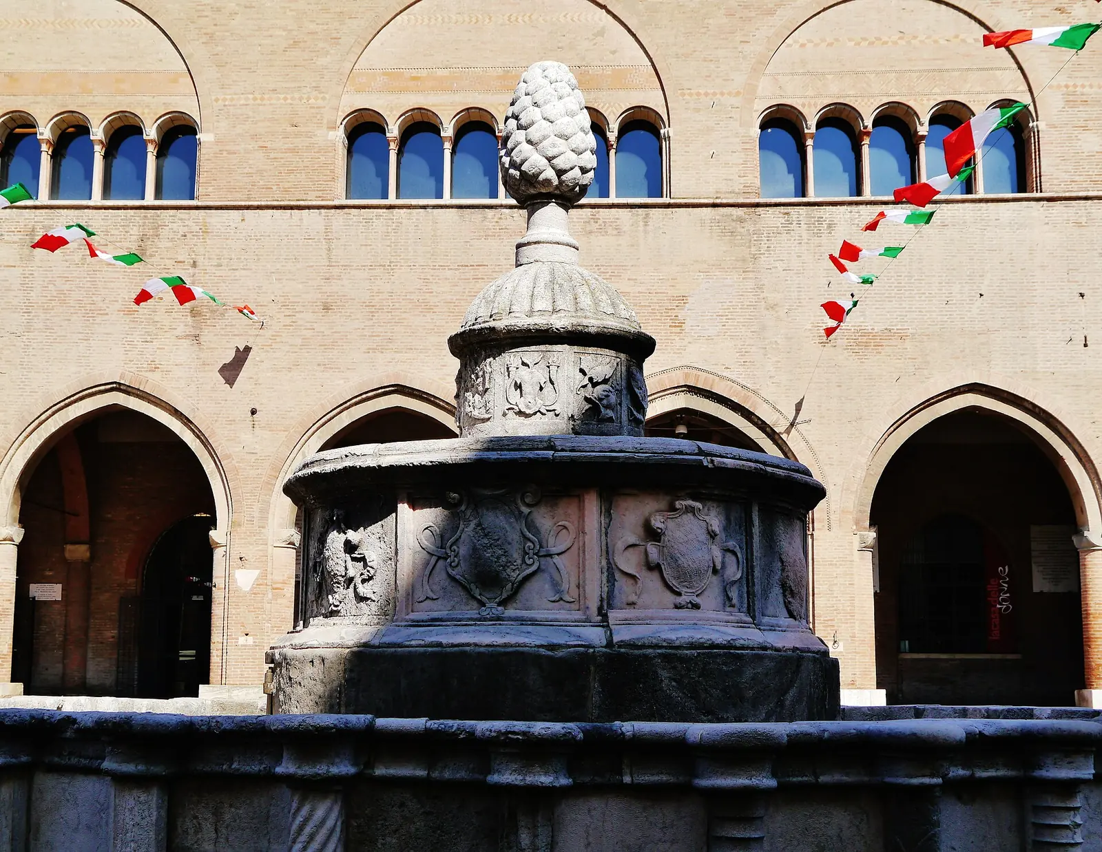The upper basin and pine cone of Fontana della Pigna in Rimini, with carved coats of arms and the medieval palace behind