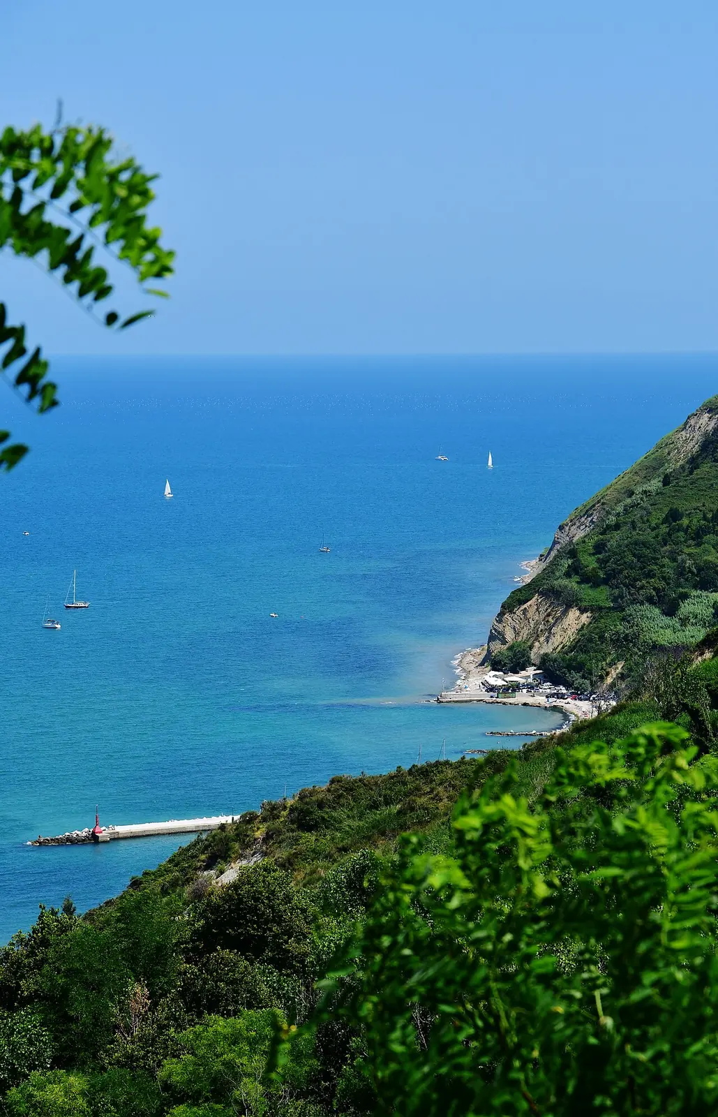 Il mare azzurro visto dall'alto di Gabicce Monte, con una piccola baia sotto la costa alta e un molo con il fanalino rosso