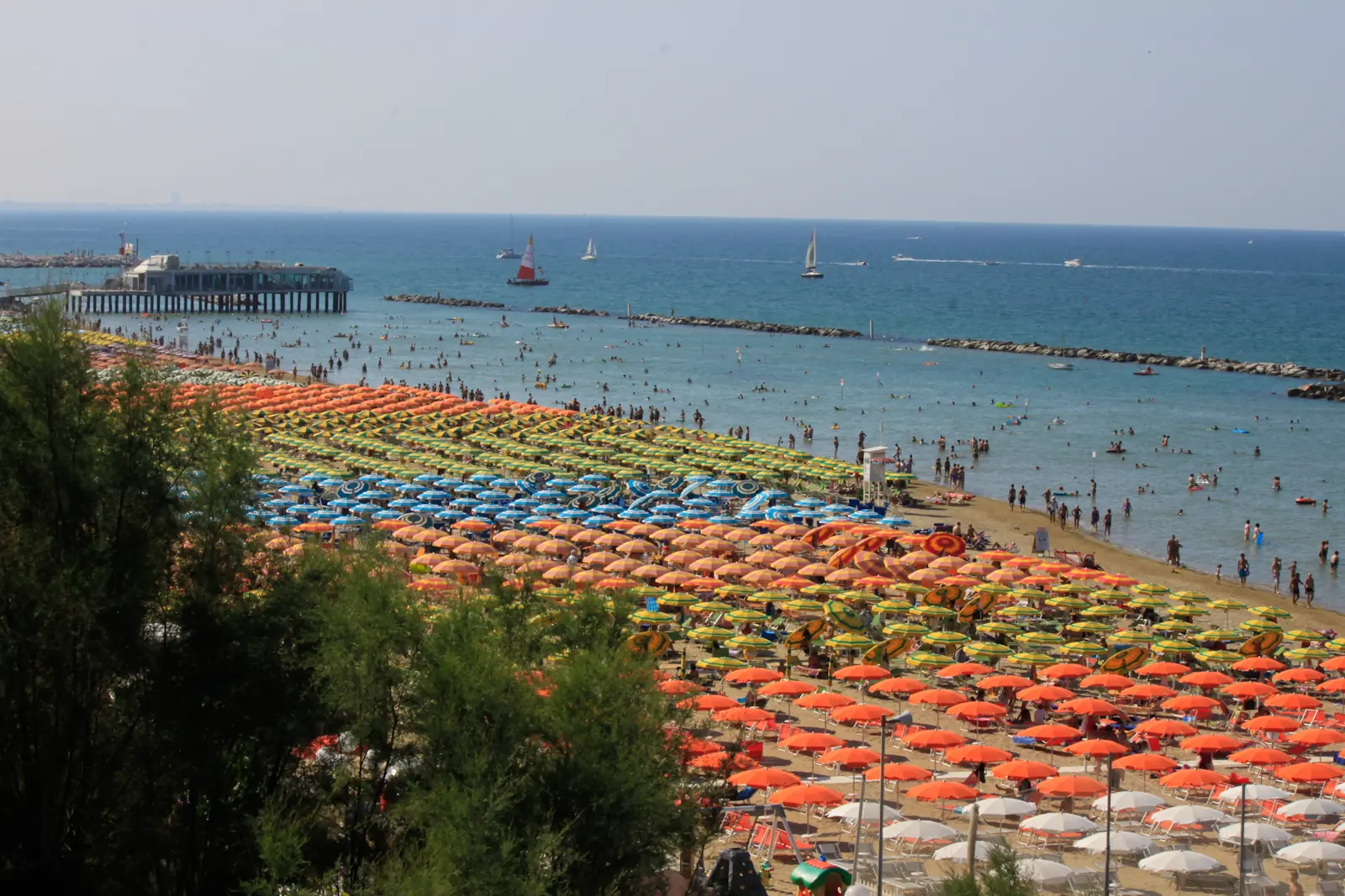 Der Strand von Gabicce Mare voller orangefarbener Sonnenschirme mit Steg, Wellenbrechern und einigen Segelbooten