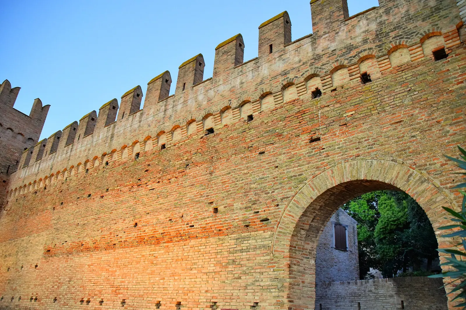 Le alte mura in laterizio di Gradara illuminate dal sole basso, con le merlature e un arco che si apre nel muro