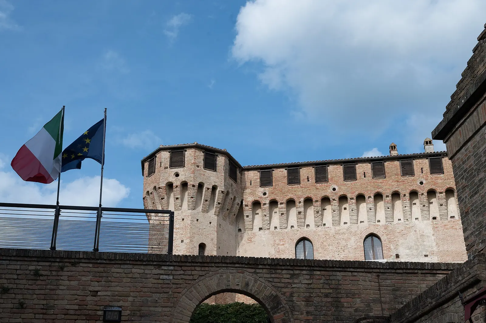 A crenellated Rocca di Gradara tower beyond a brick arch, with Italian and European flags