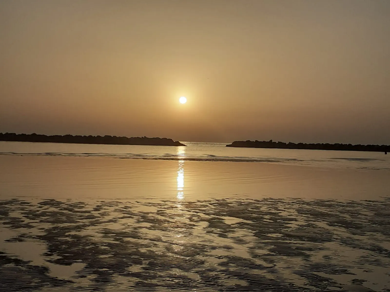 The sun above Igea Marina's calm sea, with wet sand in the foreground and two breakwaters in the distance