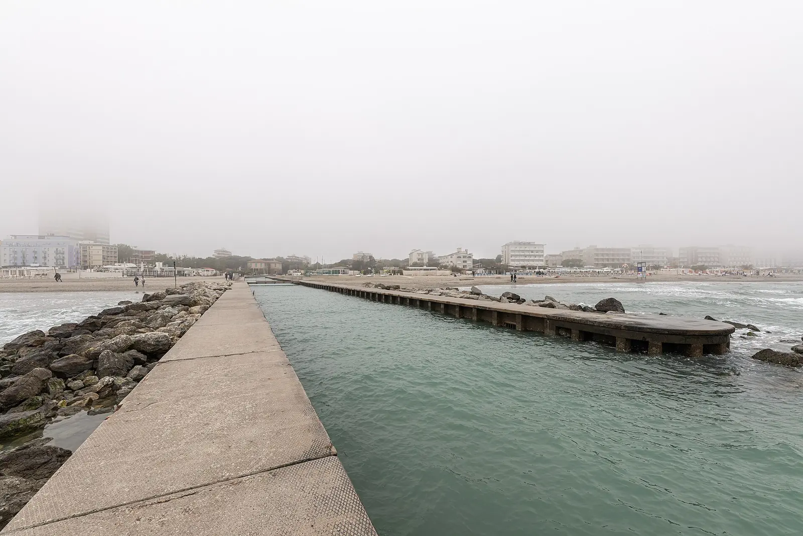 The pier and rocks at the mouth of the Canalino on a foggy day, with Milano Marittima beach in the distance