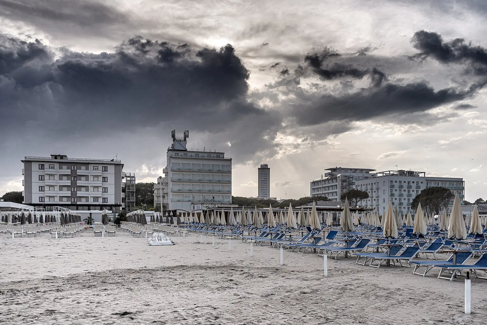 Buildings behind rows of closed parasols and blue loungers on Milano Marittima beach, beneath dark clouds