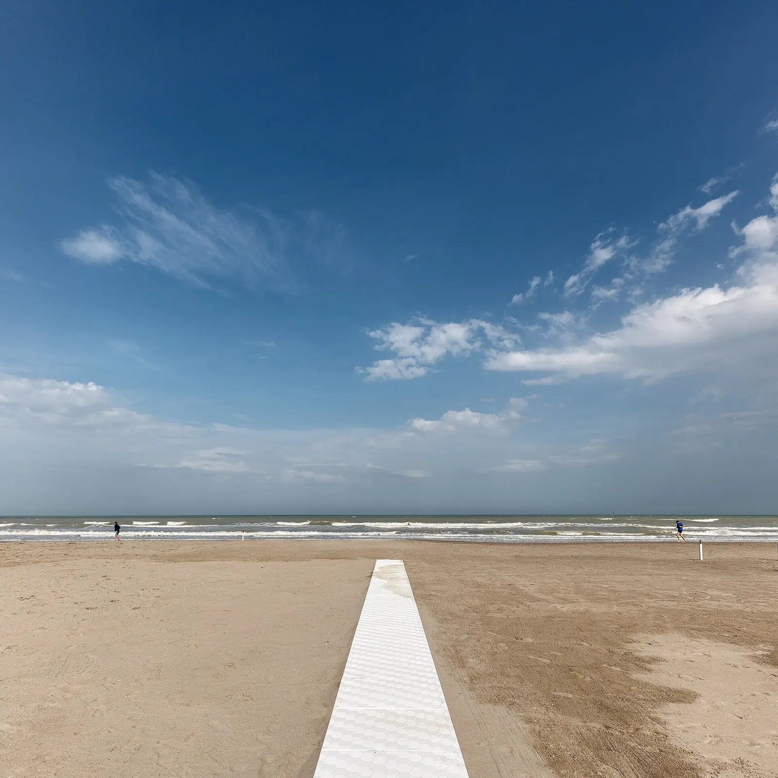 Ein weißer Steg führt über den Sand zum Meer unter weitem blauem Himmel mit wenigen Wolken