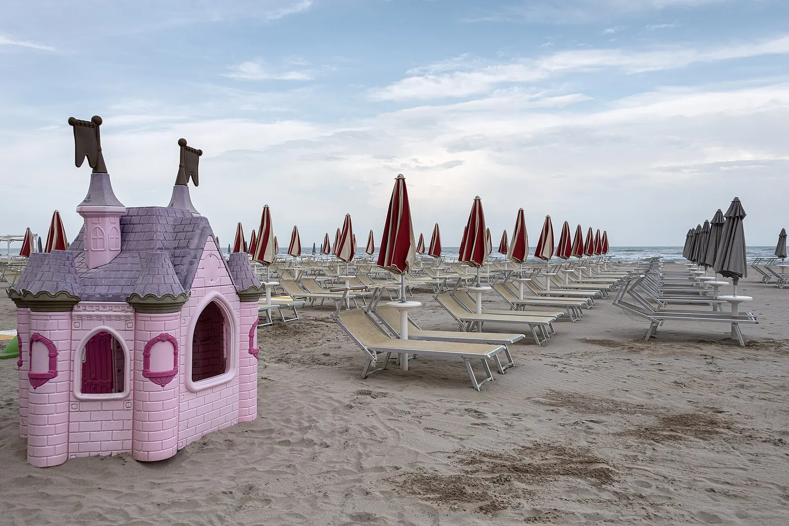 Ein rosa Spielhäuschen für Kinder im Strandsand, vor Reihen von Liegen und geschlossenen Sonnenschirmen
