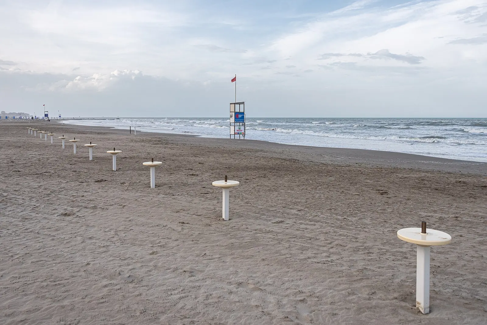 Una torretta di salvataggio con la bandiera rossa sulla spiaggia vuota, vicino a una fila di basi per ombrelloni