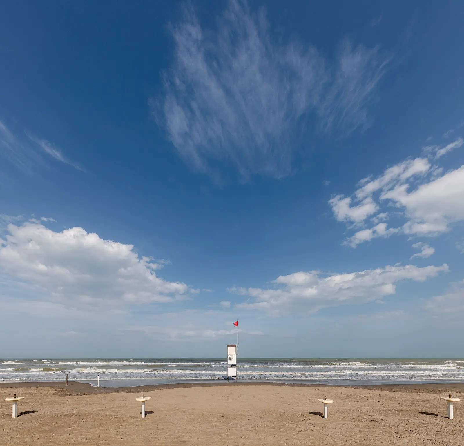 A lifeguard tower with a red flag on the shore, facing a rough sea and a blue sky with thin clouds