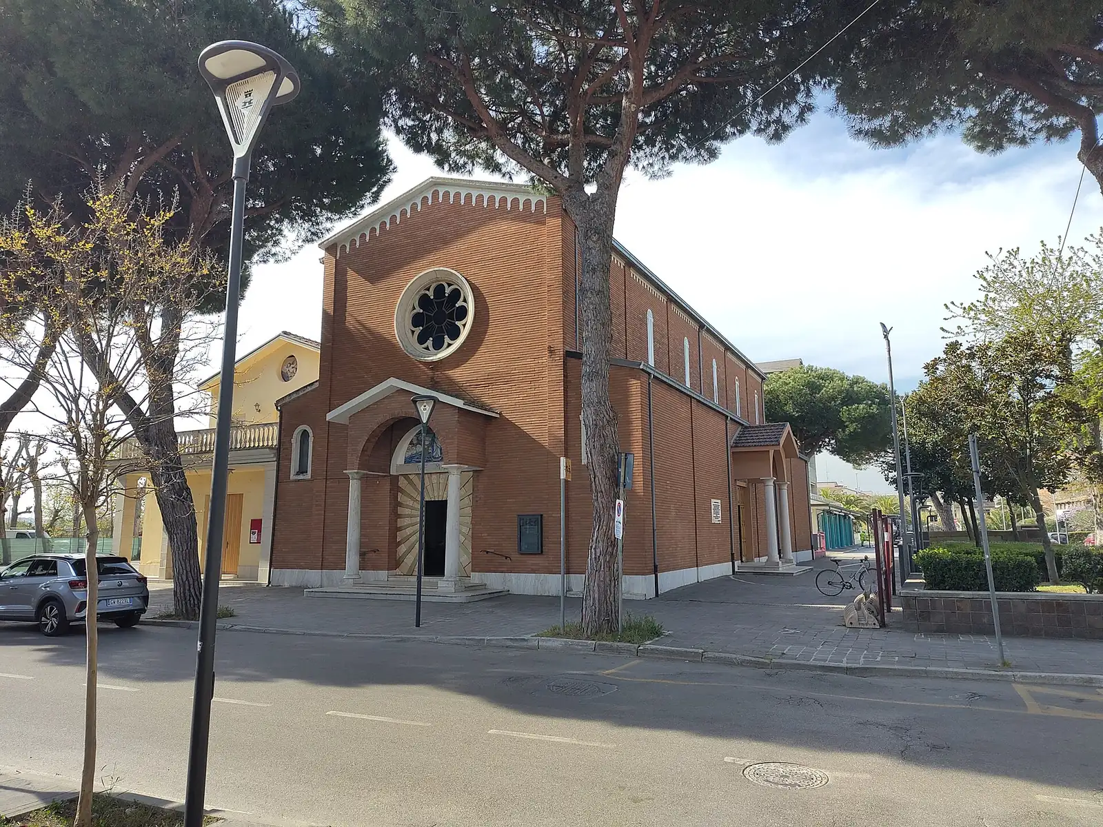 Die Backsteinfassade einer Kirche mit Rosette und Vorhalle zwischen Pinien und einer Straße in Misano Adriatico