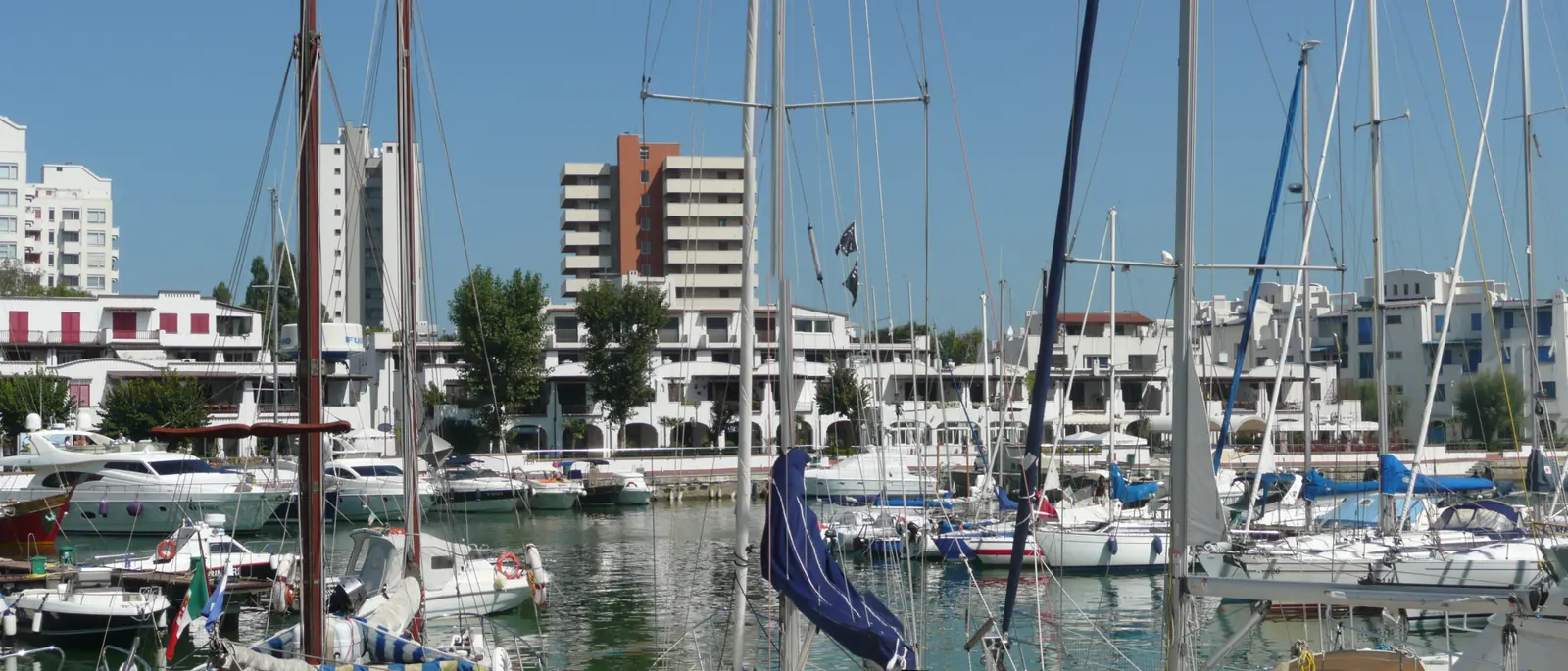 Das Hafenbecken von Portoverde mit vertäuten Segel- und Motorbooten und weißen Gebäuden mit Arkaden im Hintergrund