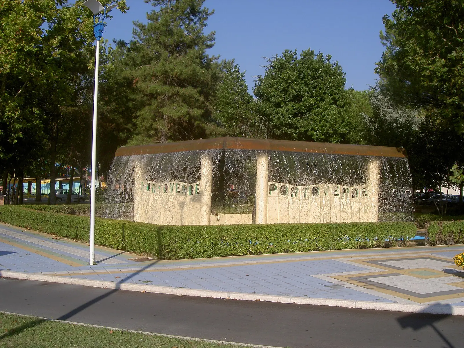 Ein Kaskadenbrunnen mit der Aufschrift Portoverde in einem Heckenbeet zwischen Bäumen und einem gemusterten Gehweg