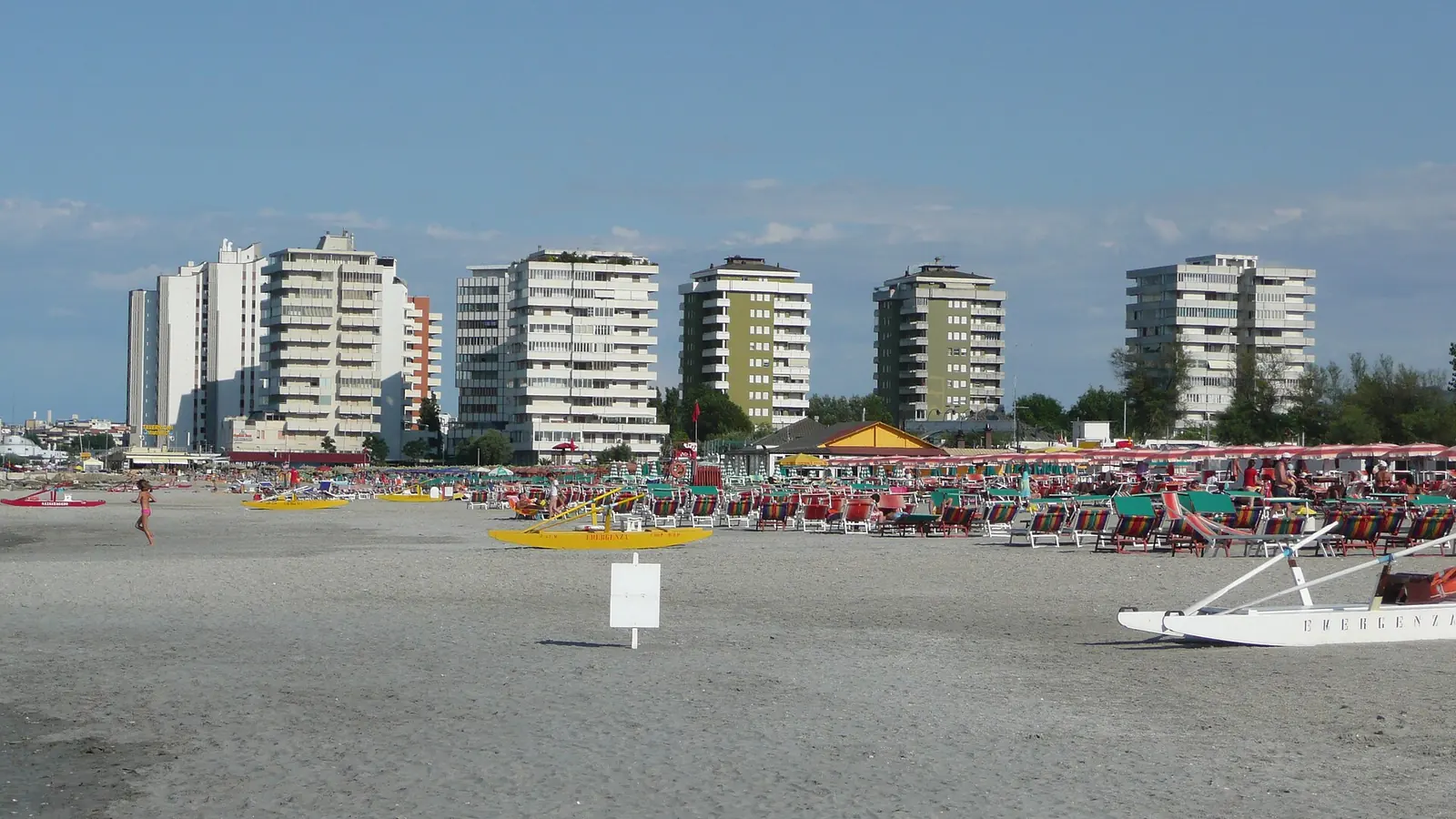 Der Strand von Portoverde mit Sonnenschirmen und Liegestühlen, einem Tretboot im Sand und hohen Gebäuden im Hintergrund