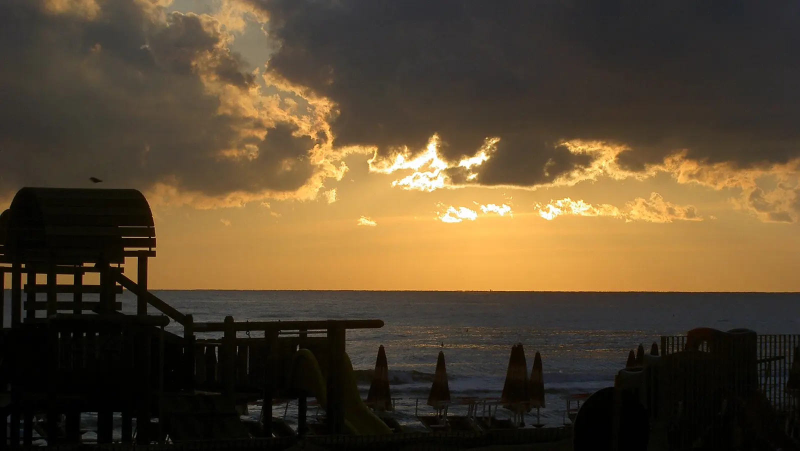 Golden clouds over the sea at sunset, with the dark silhouettes of a wooden play structure and closed parasols