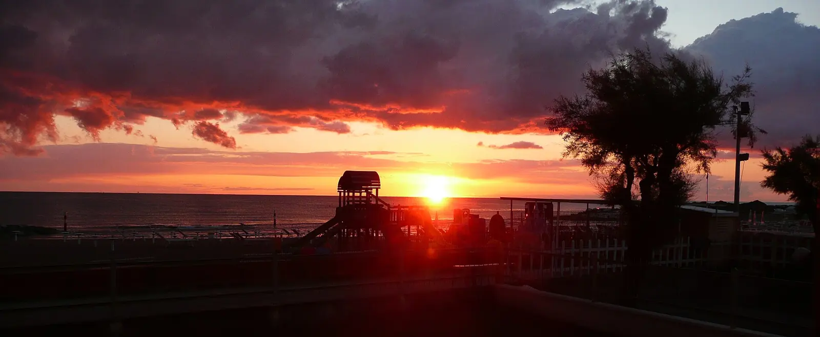 Die rote untergehende Sonne unter dunklen Wolken mit Silhouetten eines Holzspielgeräts und einer Tamariske