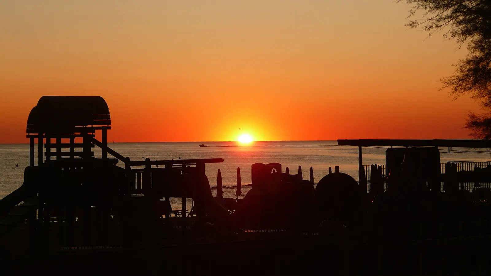 Sonnenuntergang über der Adria mit den Silhouetten eines hölzernen Kinderspielgeräts und eines Baums im Gegenlicht