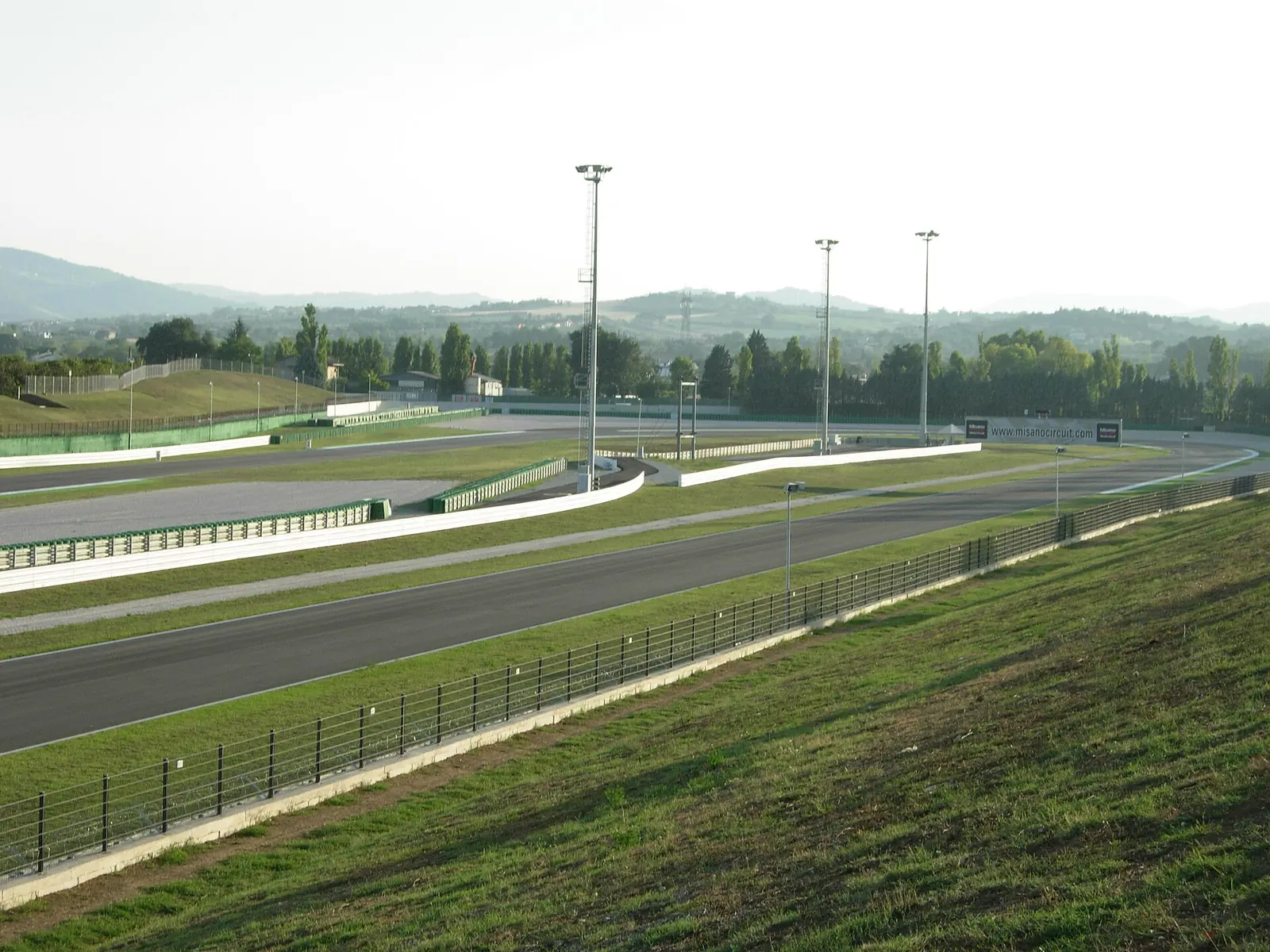 Die Gerade und eine Kurve des Misano World Circuit zwischen Wiesen und Baumreihen mit Hügeln am Horizont