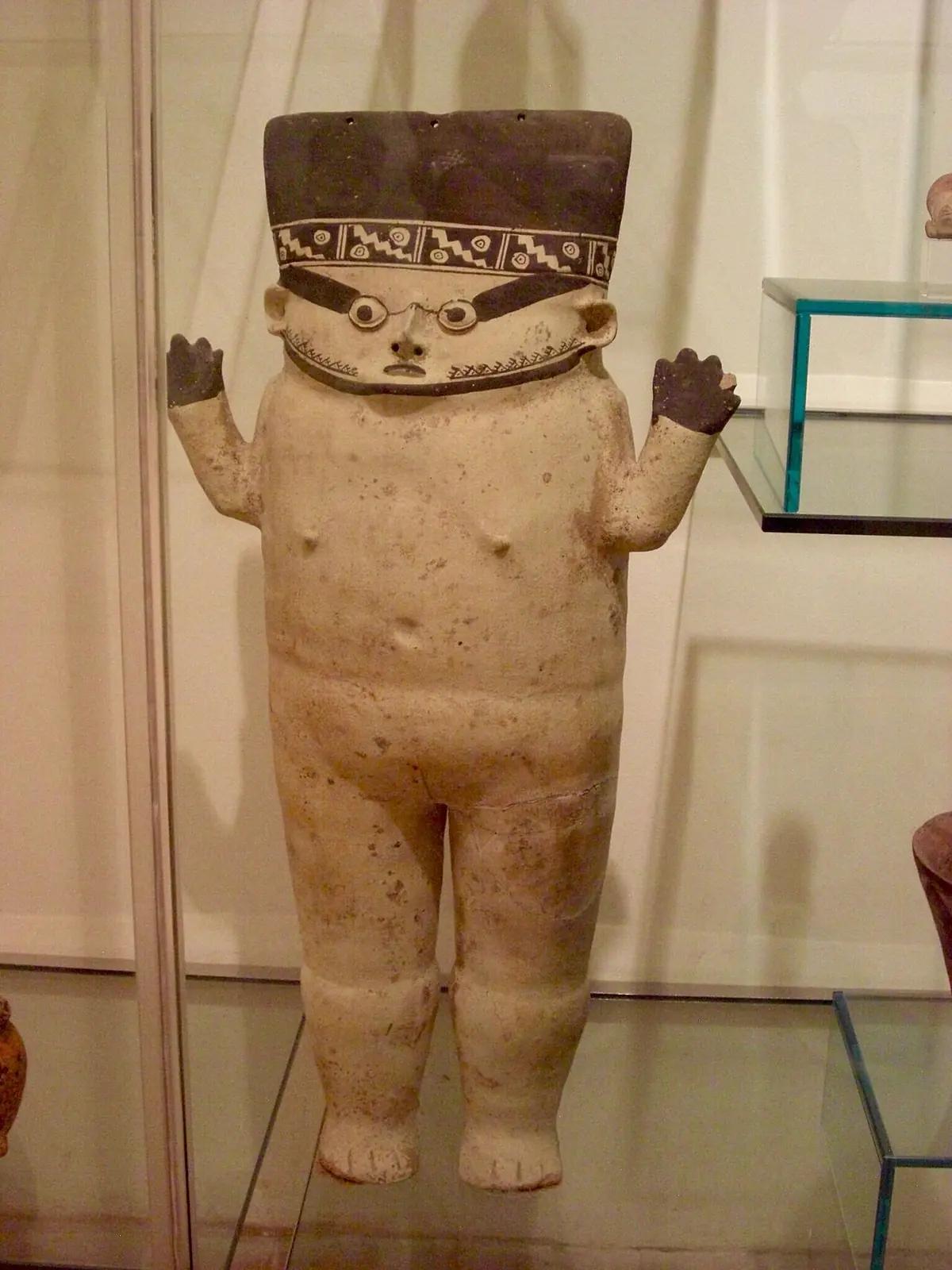 Eine große Figur aus heller Keramik mit verziertem Kopfschmuck und erhobenen Armen in einer Vitrine des Museo degli Sguardi