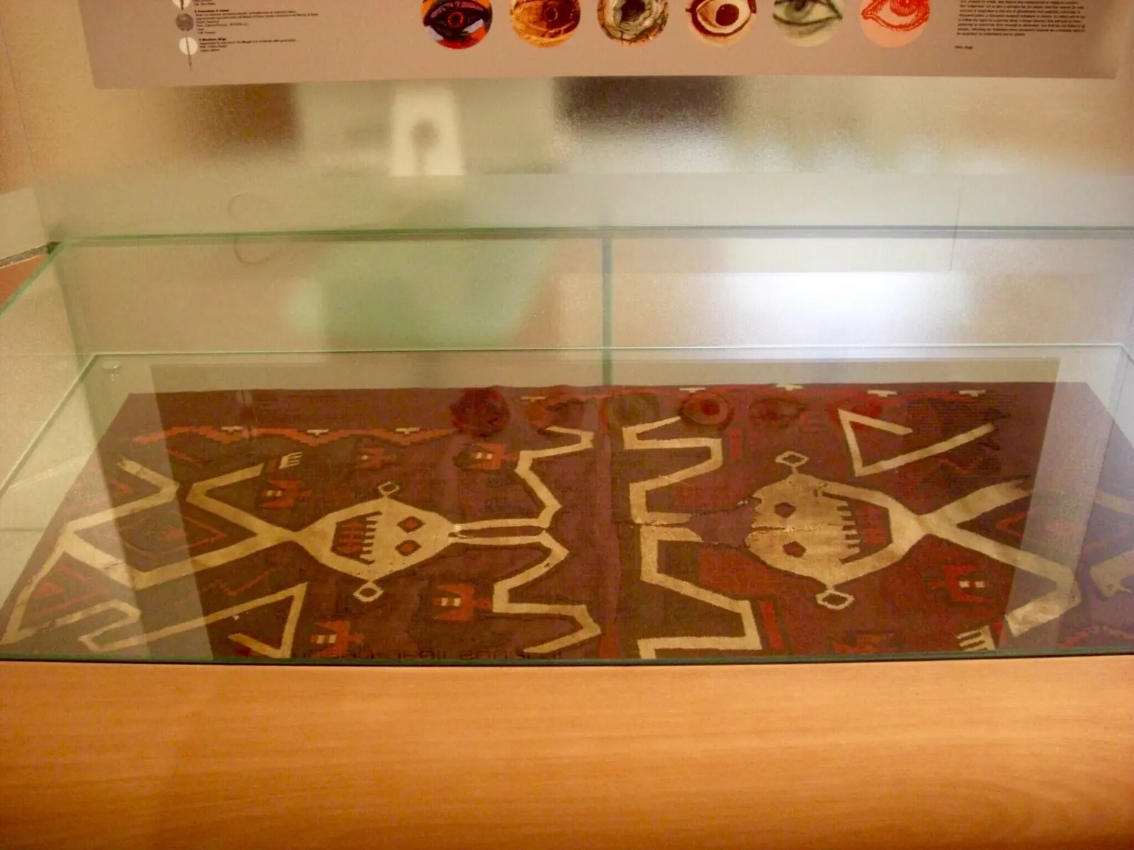 Ein altes Gewebe mit cremefarbenen geometrischen Mustern auf bordeauxrotem Grund in einer waagerechten Vitrine des Museo degli Sguardi