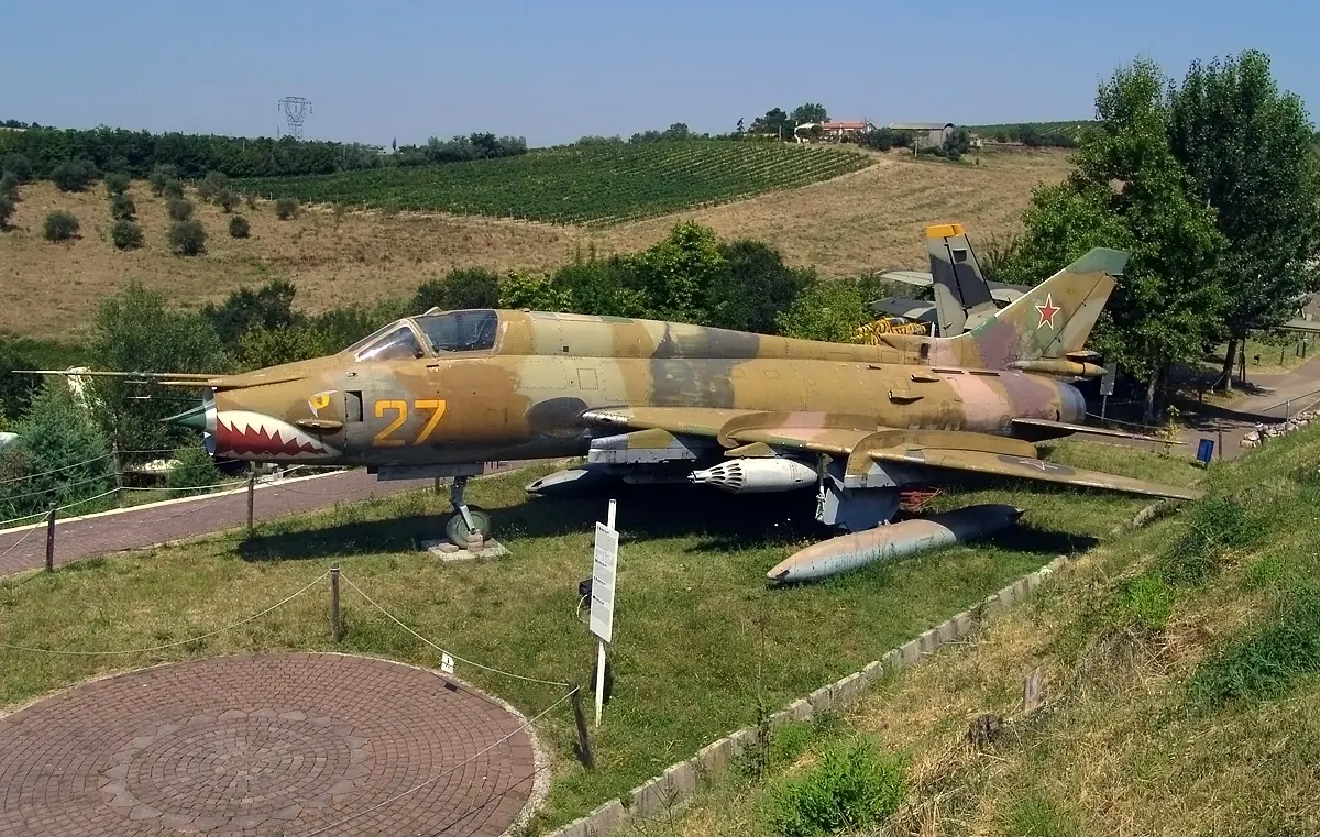 A camouflaged fighter-bomber with a shark's mouth painted on its nose, on a lawn with hills and vineyards behind
