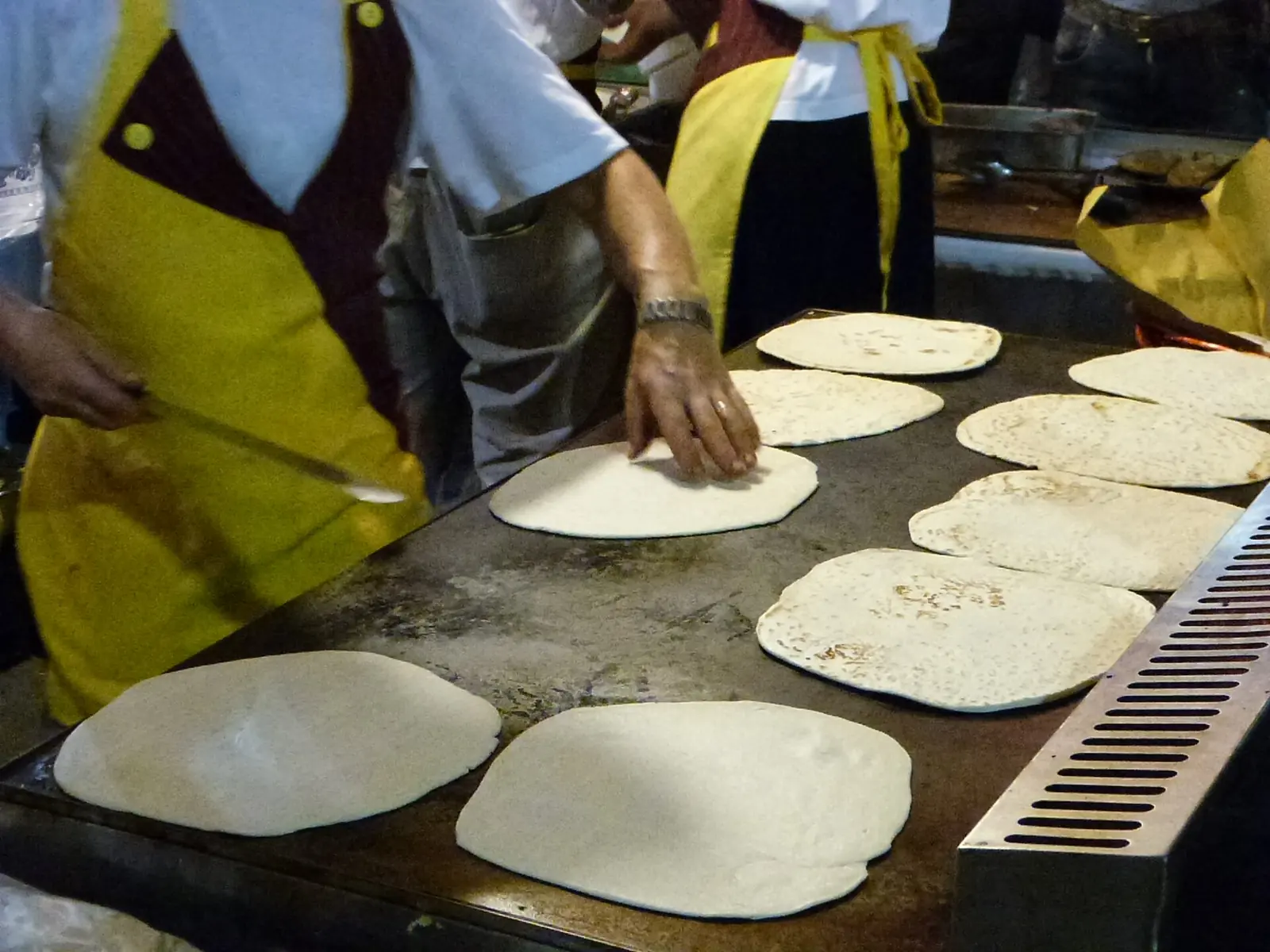 Piadine stese sulla piastra durante la cottura, con due cuochi in grembiule giallo di cui non si vedono i volti