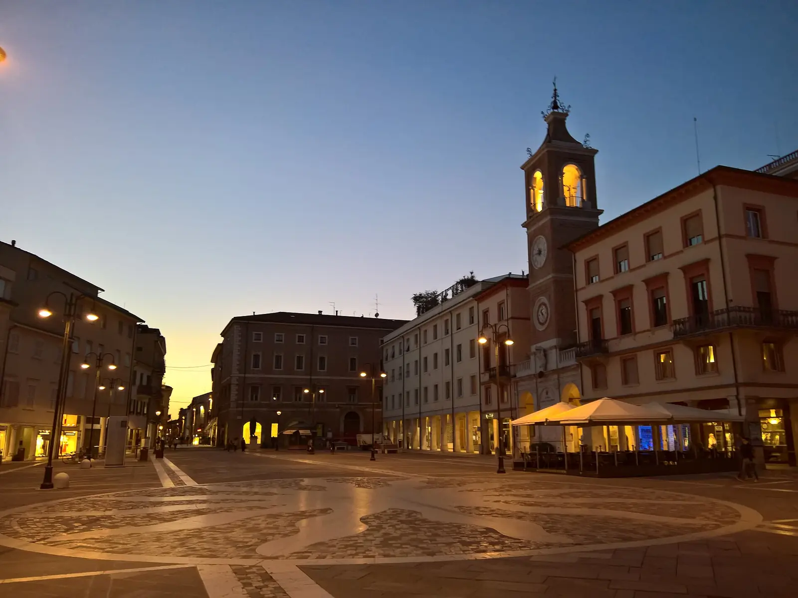 Piazza Tre Martiri a Rimini al crepuscolo, con la Torre dell'Orologio illuminata e il disegno a mosaico del pavimento