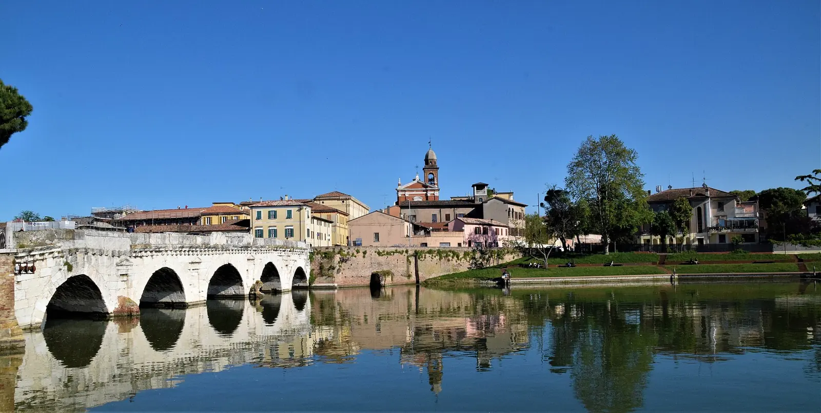 Le Ponte di Tiberio à Rimini avec ses arches reflétées dans l'eau et les maisons du bourg en arrière-plan sous un ciel bleu