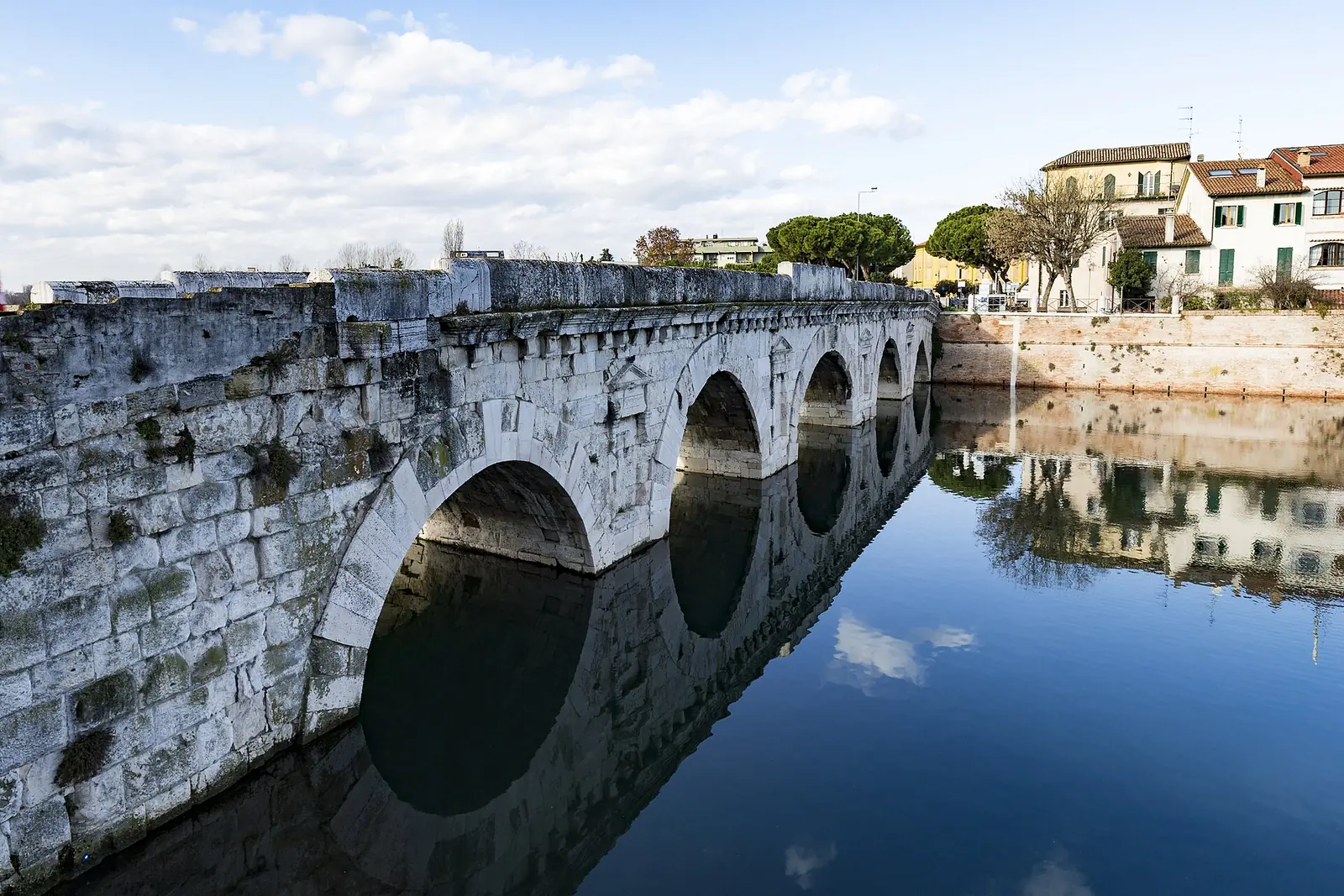 Le flanc en pierre du Ponte di Tiberio à Rimini, avec les arches et la corniche reflétées dans l'eau bleue